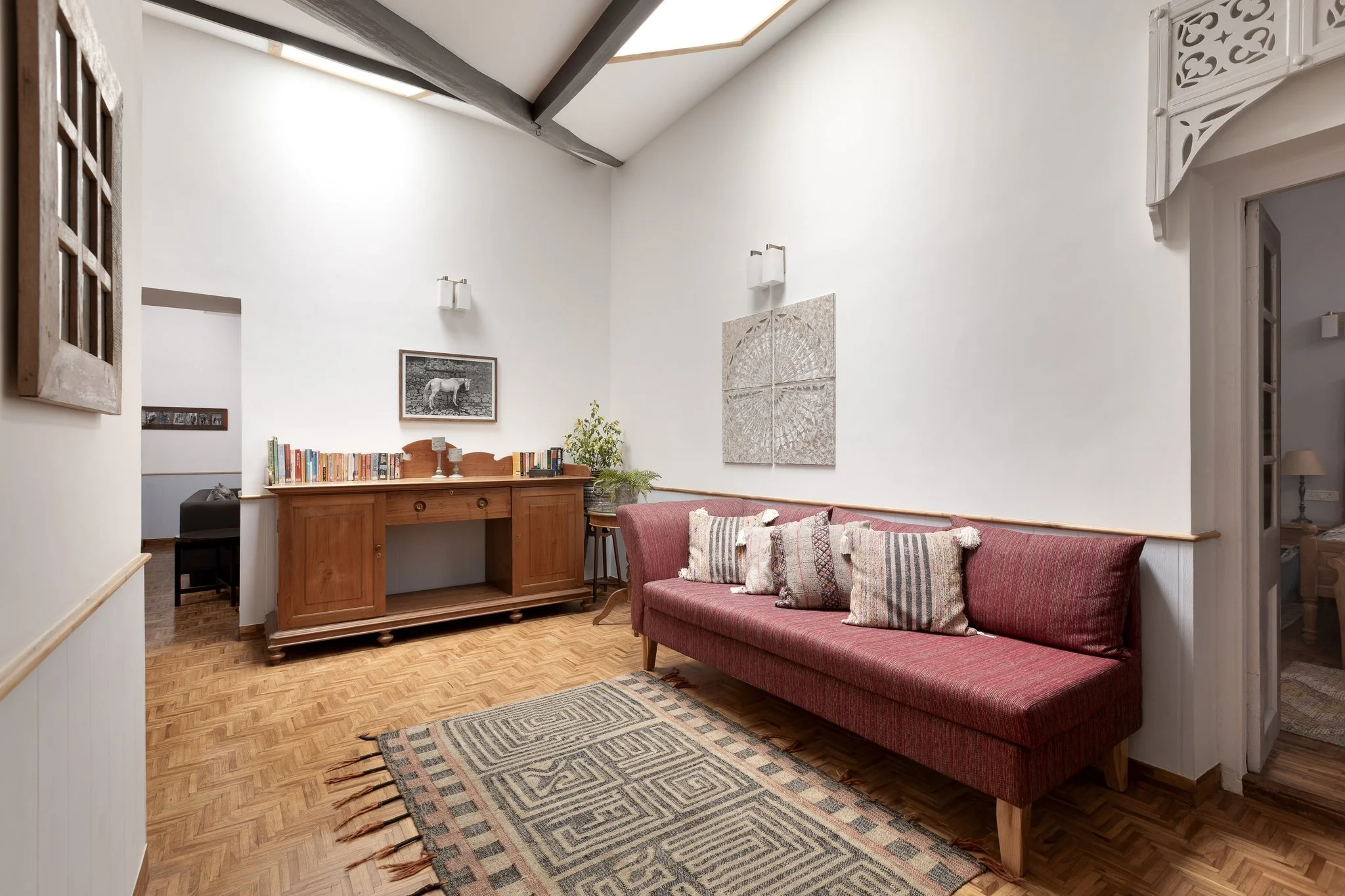 A living room with a high vaulted ceiling and wooden beams. There is a pink upholstered sofa with decorative pillows, a patterned rug on a wooden floor, a wooden sideboard with books and a small lamp, and framed artwork on the walls. A plant is place
