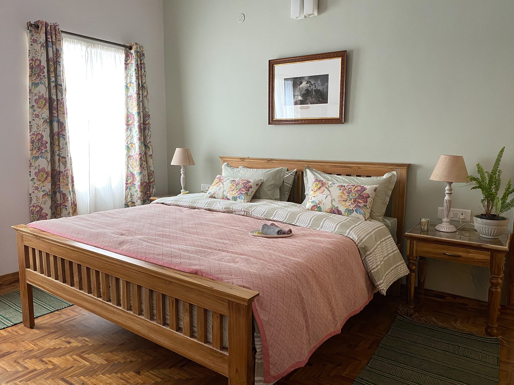 A neatly made bed with a wooden frame, floral pillows, and a pink quilt, with two nightstands on either side holding lamps, a potted plant, and a framed picture on the wall.