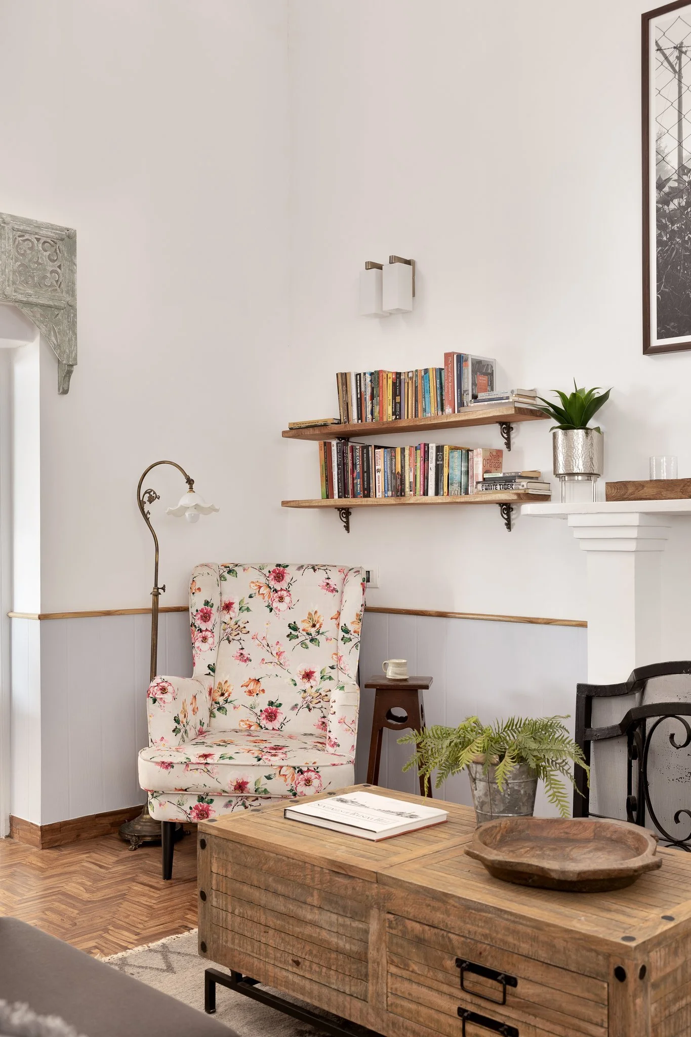 A cozy interior living room area featuring a floral upholstered armchair, a wooden coffee table with a plant and wooden tray, a small side table with a cup, bookshelf shelves filled with books on the wall, a modern wall sconce, and part of a fireplac