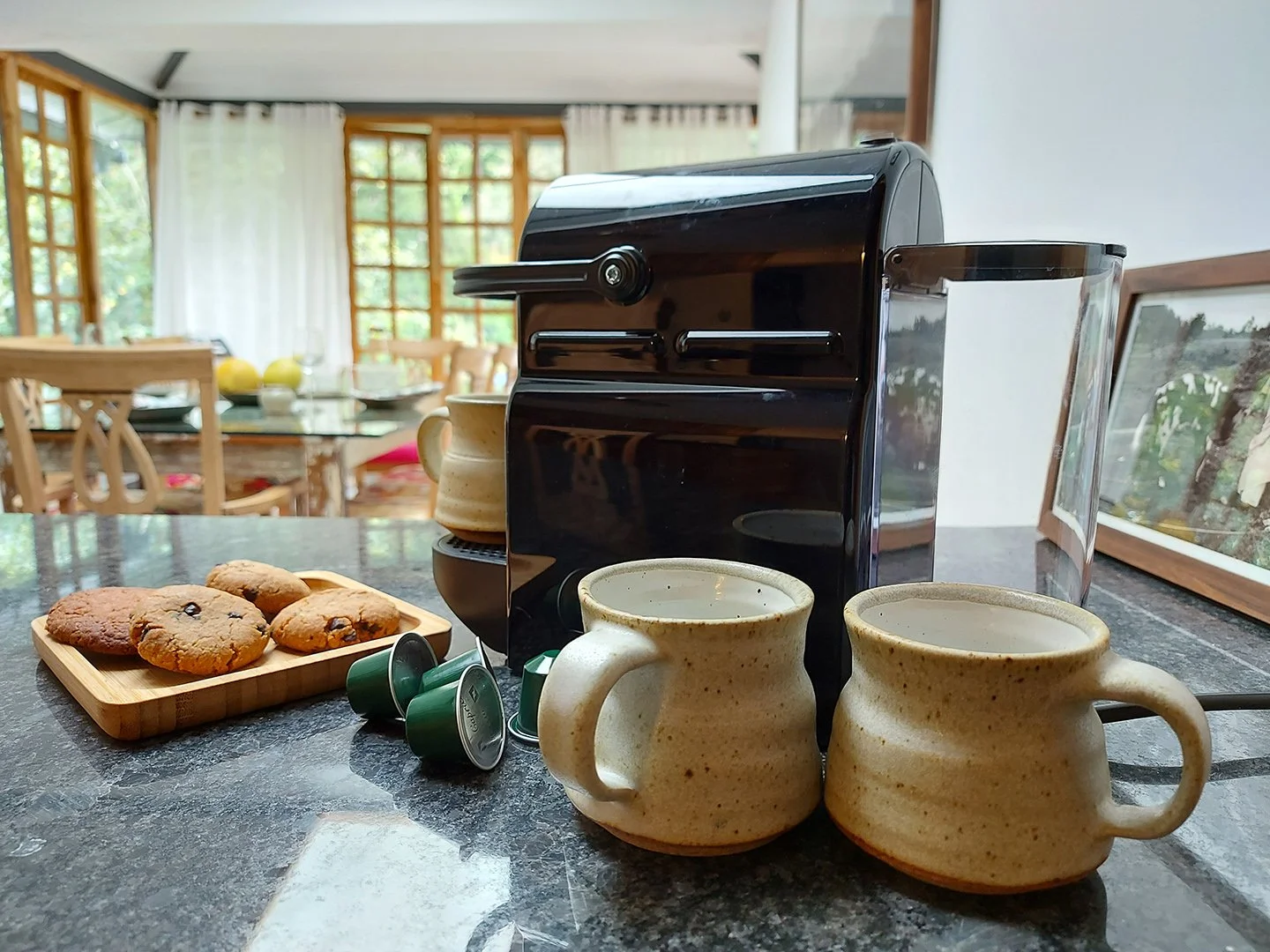 A coffee station with a black espresso machine, two ceramic mugs filled with coffee, a small wooden tray with chocolate chip cookies, and three green coffee pods on a dark, polished countertop in a bright room with large windows and wooden furniture 