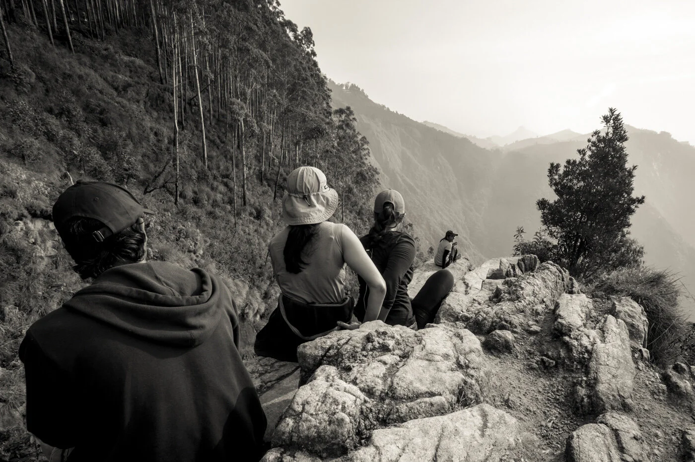 Four hikers sitting on rocky terrain on a mountain trail, overlooking a valley with trees and foggy hills in the distance.