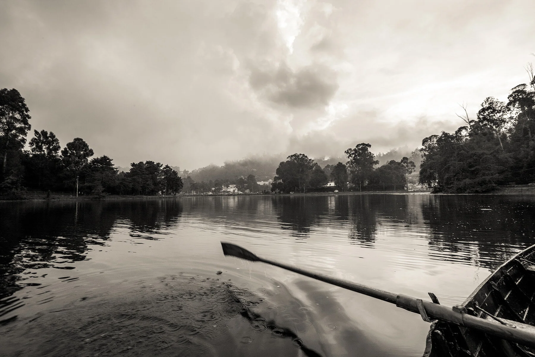 A black and white photo of a lake surrounded by trees with a cloudy sky overhead. A kayak paddle is visible in the bottom right corner of the image.