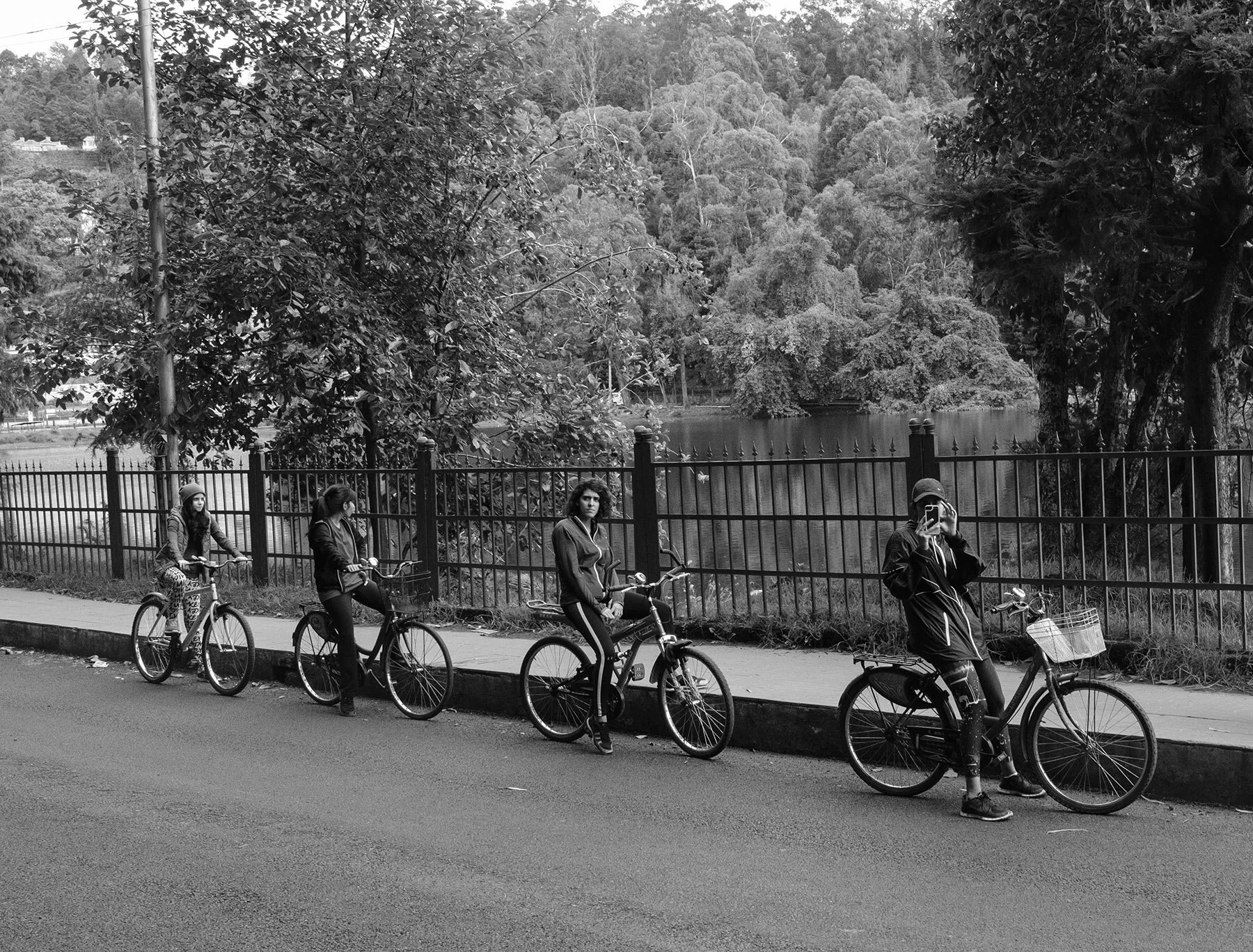 Five people with bicycles standing or riding along a sidewalk next to a black metal fence, with trees and a lake in the background.