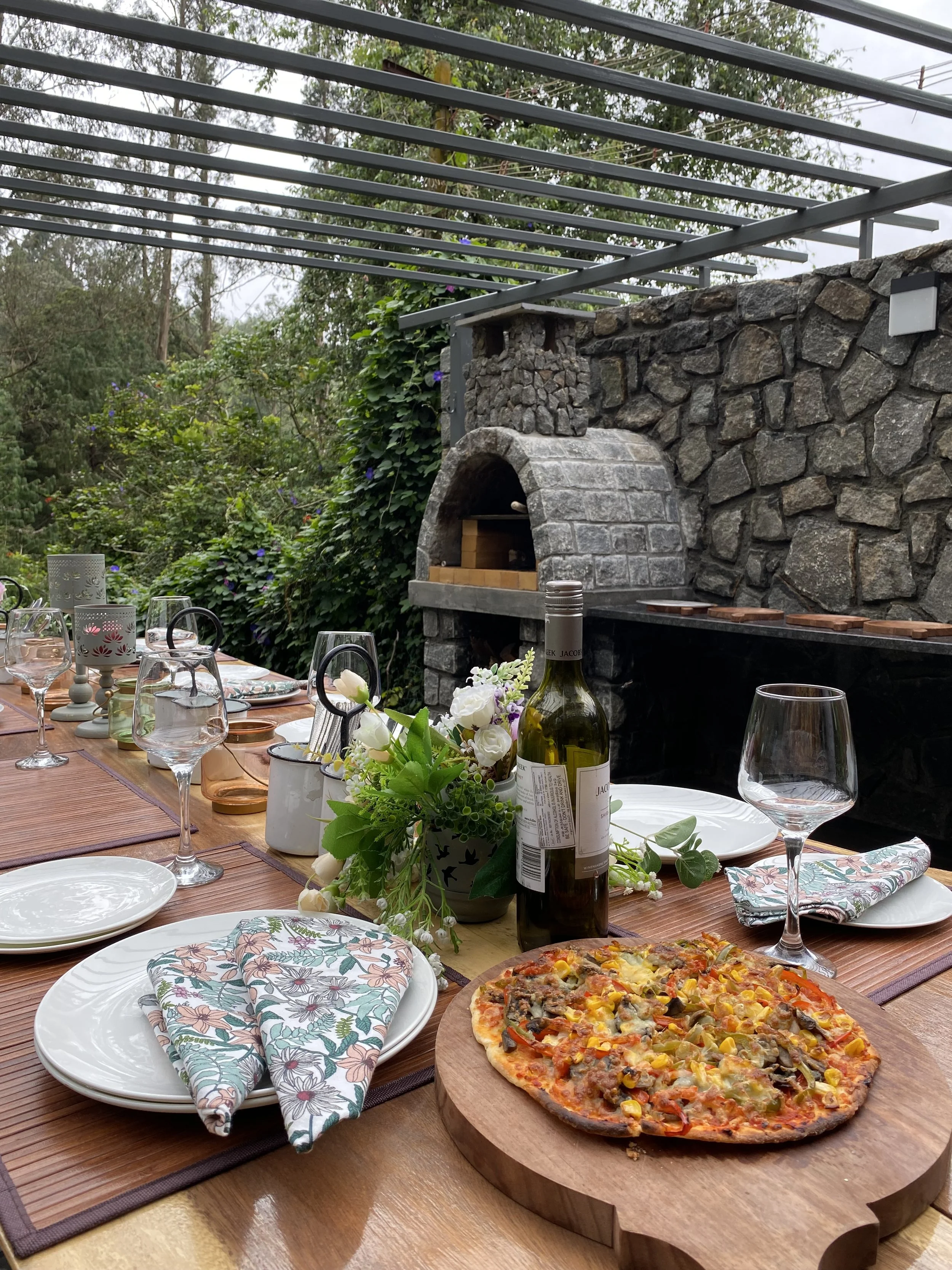 A dining table set outdoors with plates, glasses, and napkins, a flower centerpiece, a pizza on a wooden serving board, and a stone outdoor grill in the background.