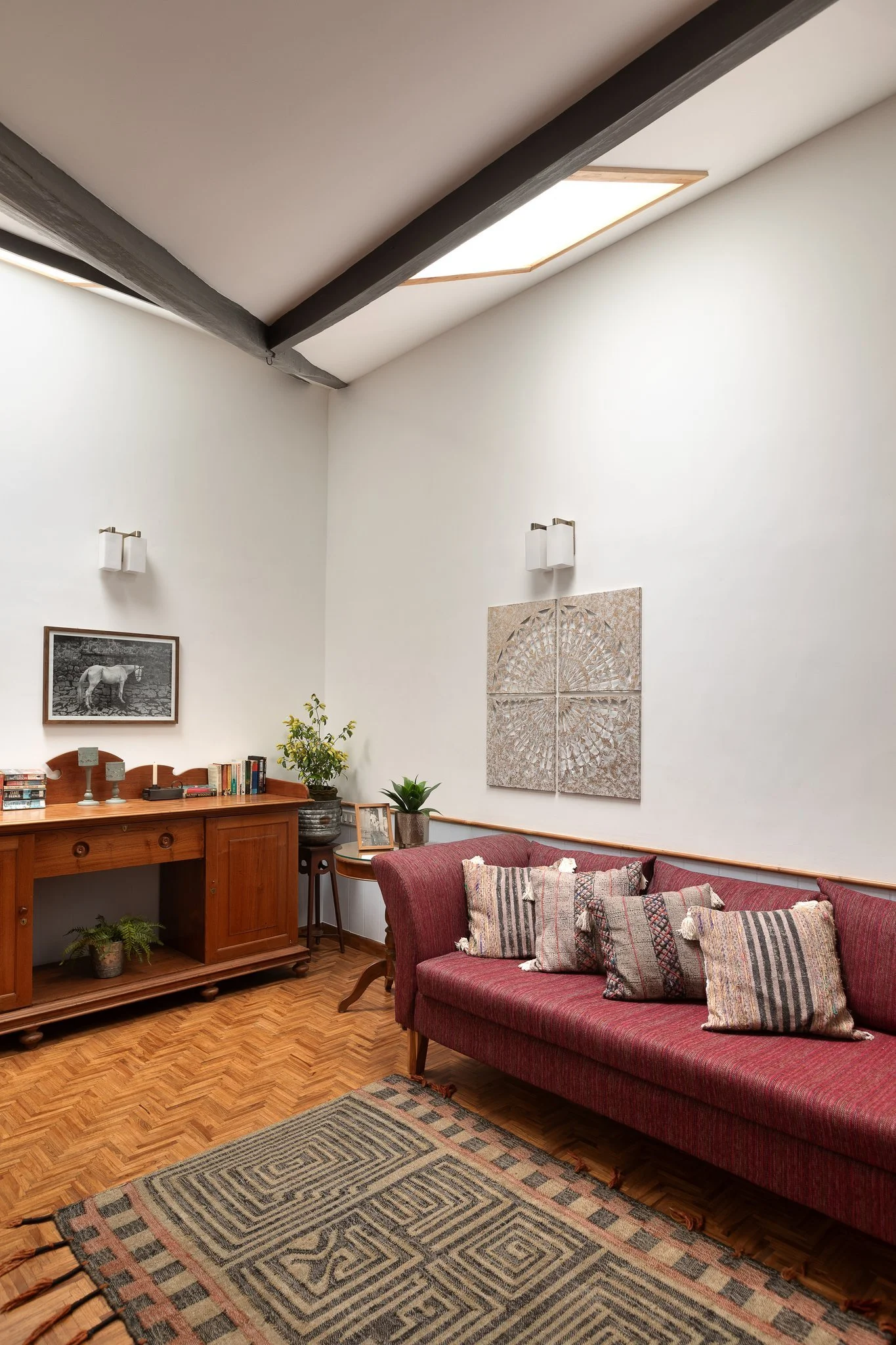 A cozy living room corner with a red upholstered sofa with patterned pillows, a wooden console table with books and decorative items, a potted plant, framed black and white photo of a horse on the wall, and a skylight in the ceiling illuminating the 