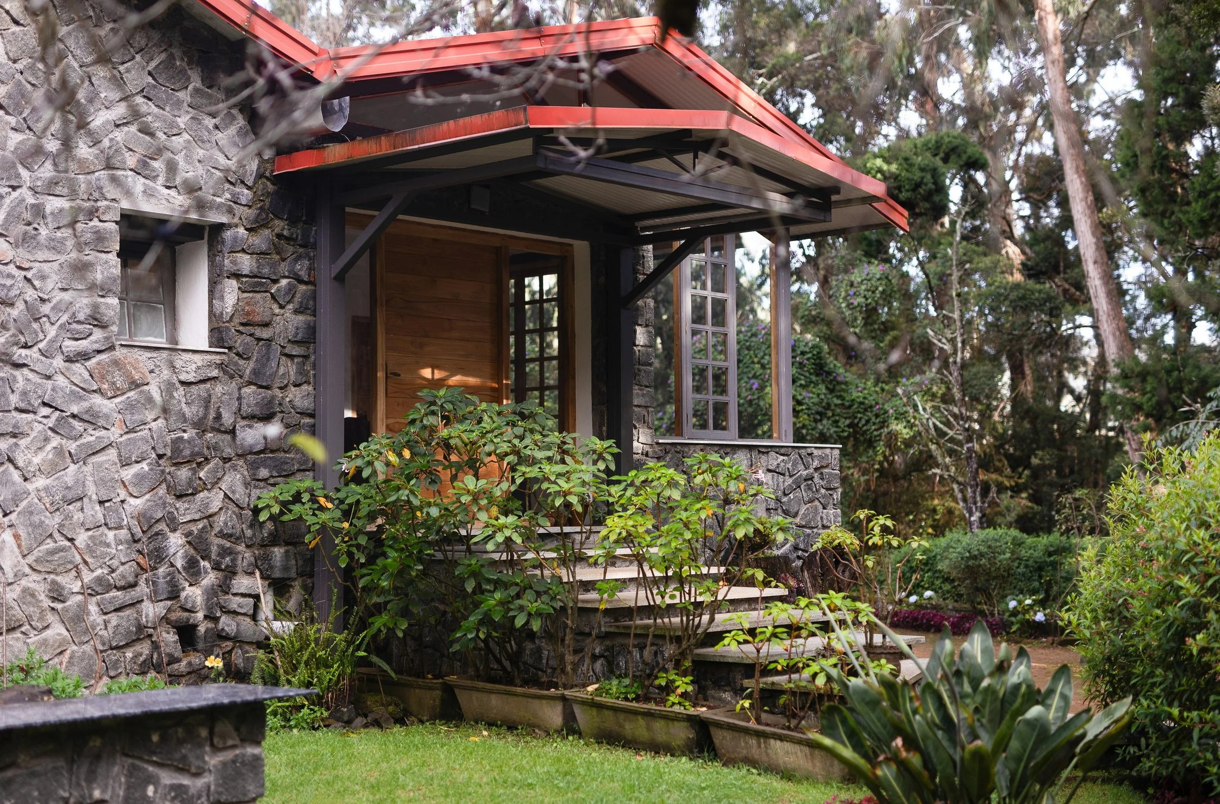 A stone house with a red metal roof and a small porch surrounded by trees and lush greenery.