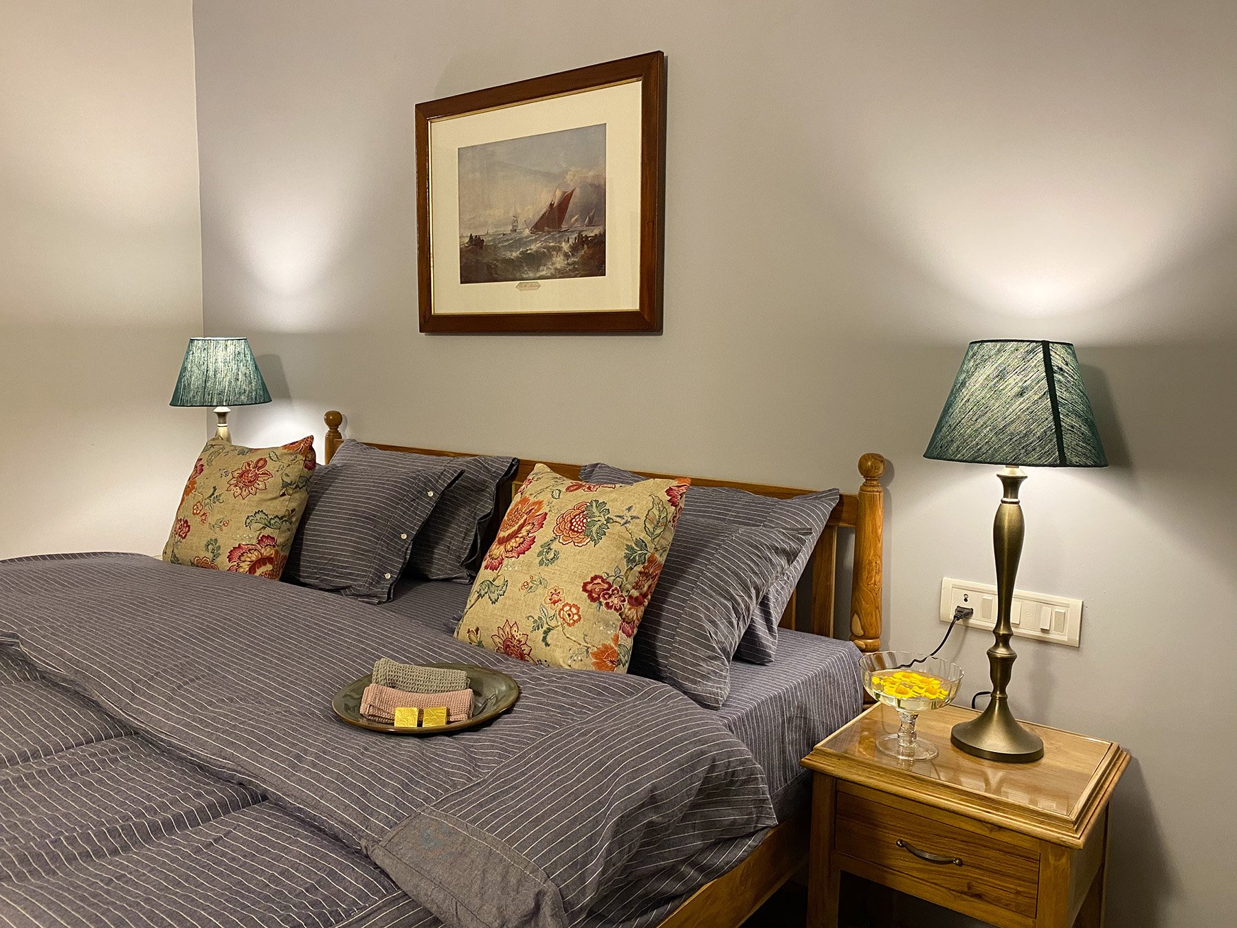 A cozy bedroom with a wooden bed frame, gray striped bedding, and multiple decorative pillows, including floral designs. There are two matching lamps with green shades on bedside tables. A framed painting of sailing ships hangs on the cream-colored w