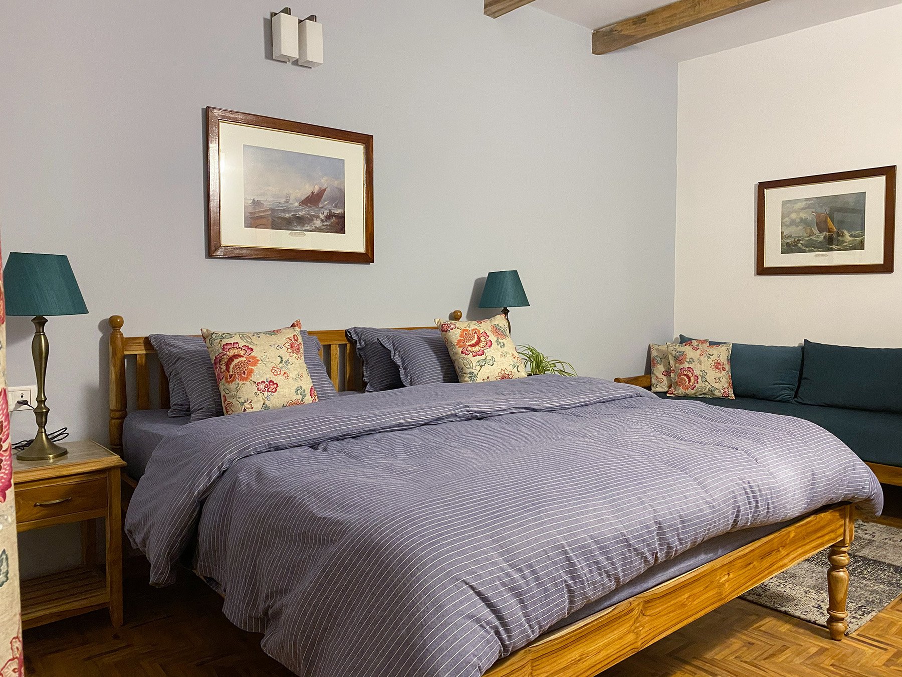 A bedroom with a wooden bed frame, a purple striped bedspread, floral and striped pillows, and side tables with teal lamps. The walls are white with two framed nautical paintings. There is wooden flooring and a visible ceiling beam.