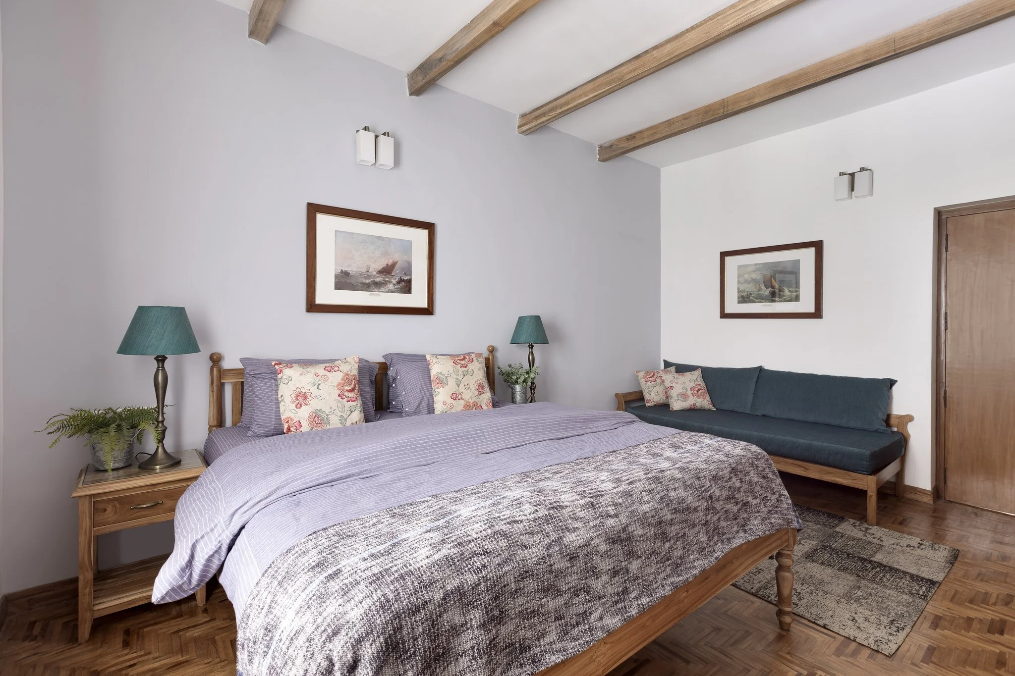 A bedroom with a wooden bed frame, light purple bedding, and four floral pillows. Two teal lamps are on a nightstand, and two framed pictures of ocean scenes hang on the light gray wall. There's a small plant on the nightstand and a carpet on the woo