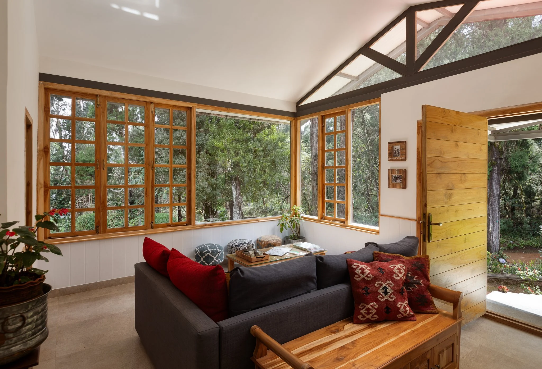 Living room with large windows showing green trees outside, wooden accents, and cozy furniture including a black sofa with red and patterned throw pillows, a small table with magazines, and potted plants.