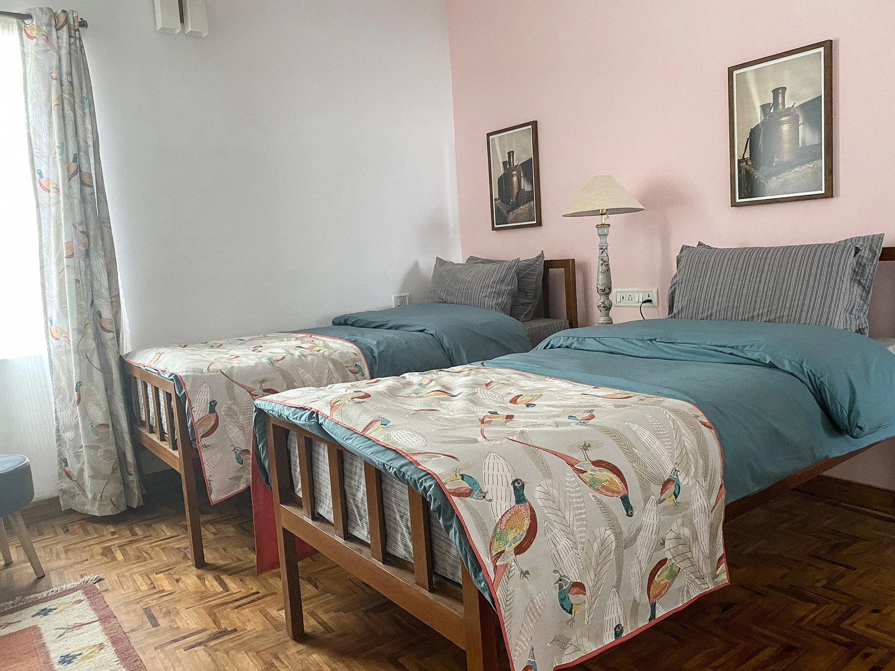A bedroom with two wooden twin beds, each covered with bedding featuring colorful birds and feathers. There are gray striped pillows on each bed, a lamp between them, and framed pictures of vintage coffee grinders on the pink and white walls. A windo