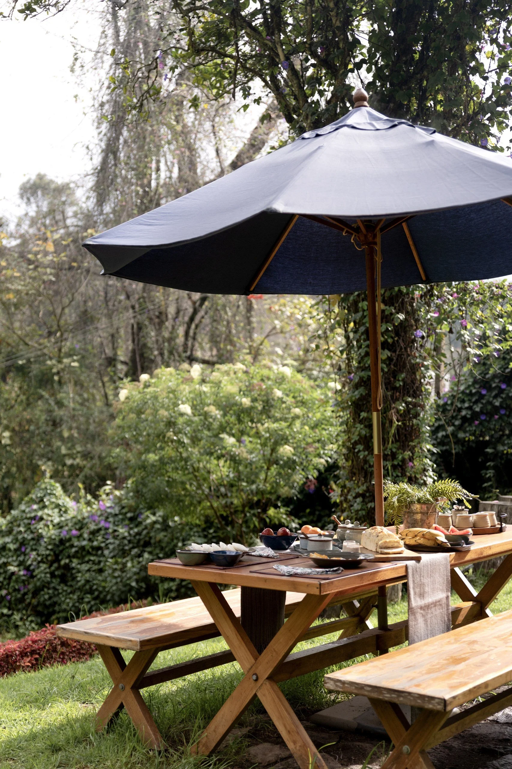 Outdoor wooden table set for a meal in a garden, shaded by a large blue patio umbrella, surrounded by lush greenery and blooming plants.