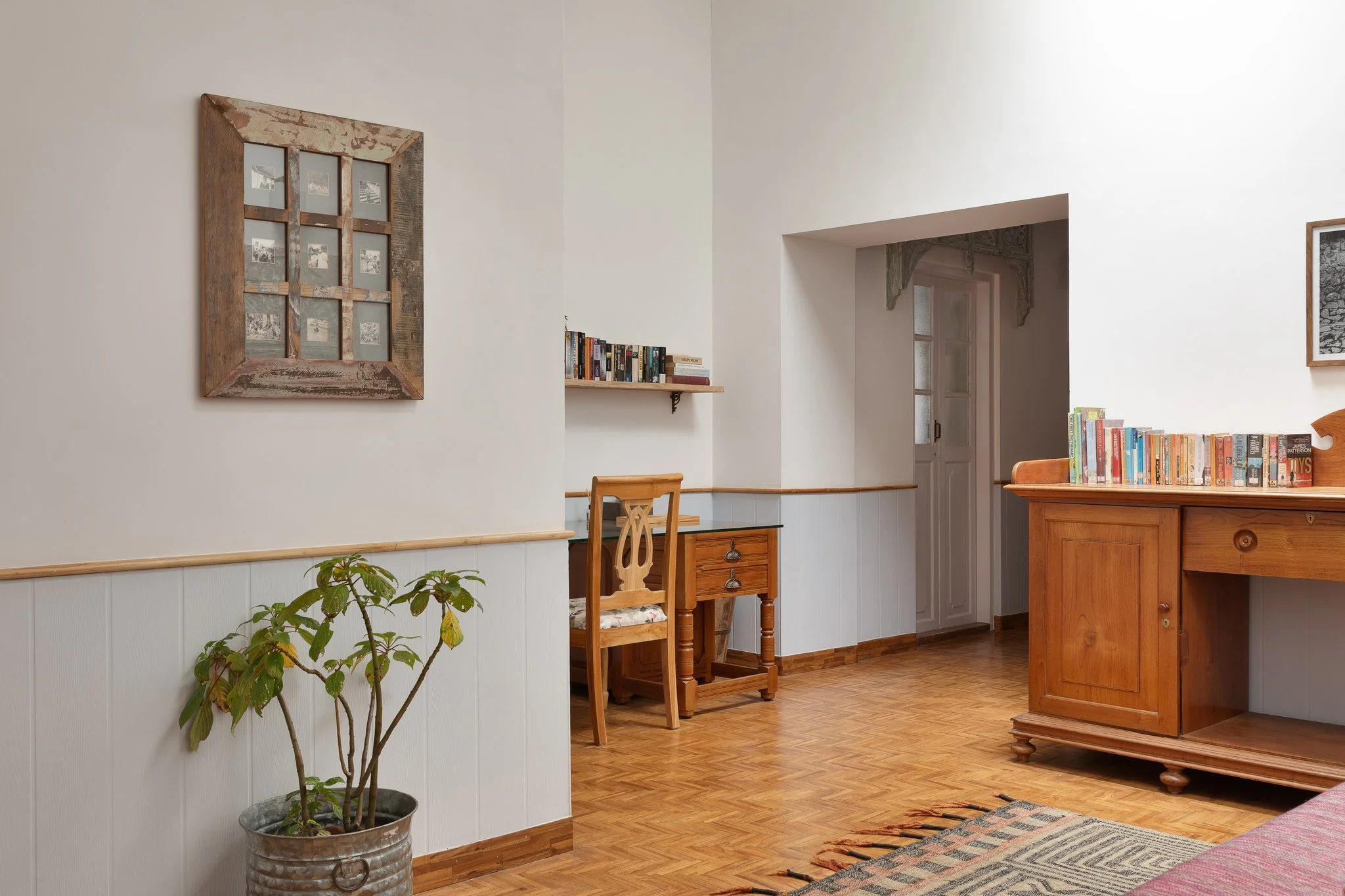 Interior of a room with white walls, wooden flooring, and a wooden table. Contains framed pictures on the walls, a bookshelf, and a potted plant in the foreground.