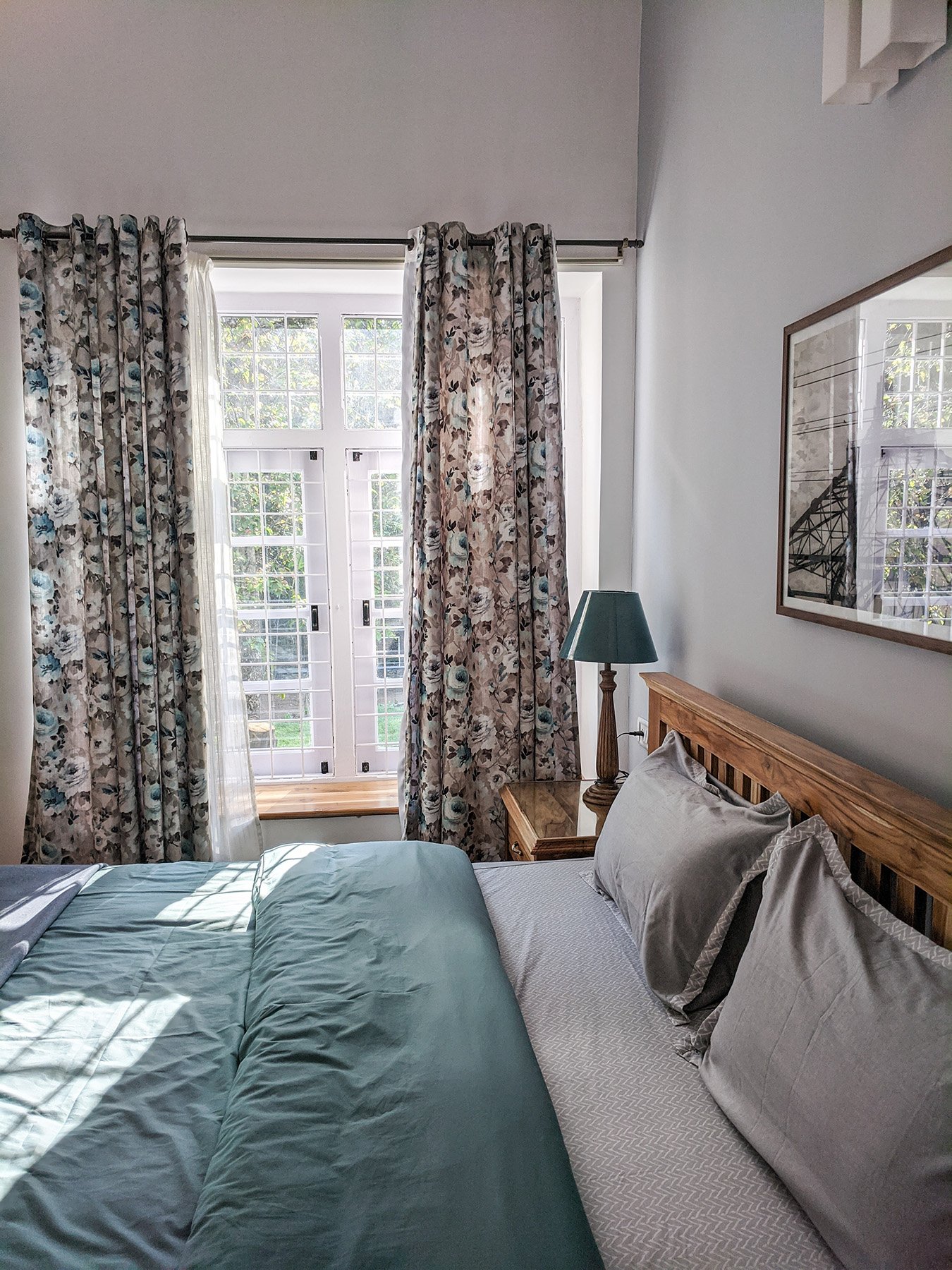A bedroom with a bed, green bedding, and pillows, a wooden nightstand with a green lampshade, floral curtains, and a window with white grid framing and lush greenery outside.