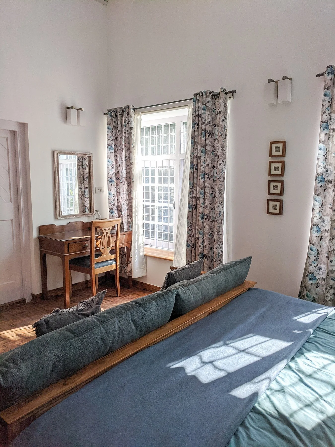 Living room with a wooden desk and chair, a window with floral curtains, a mirror on the wall, and a couch with pillows, sunlight casting shadows, and a framed artwork collection.