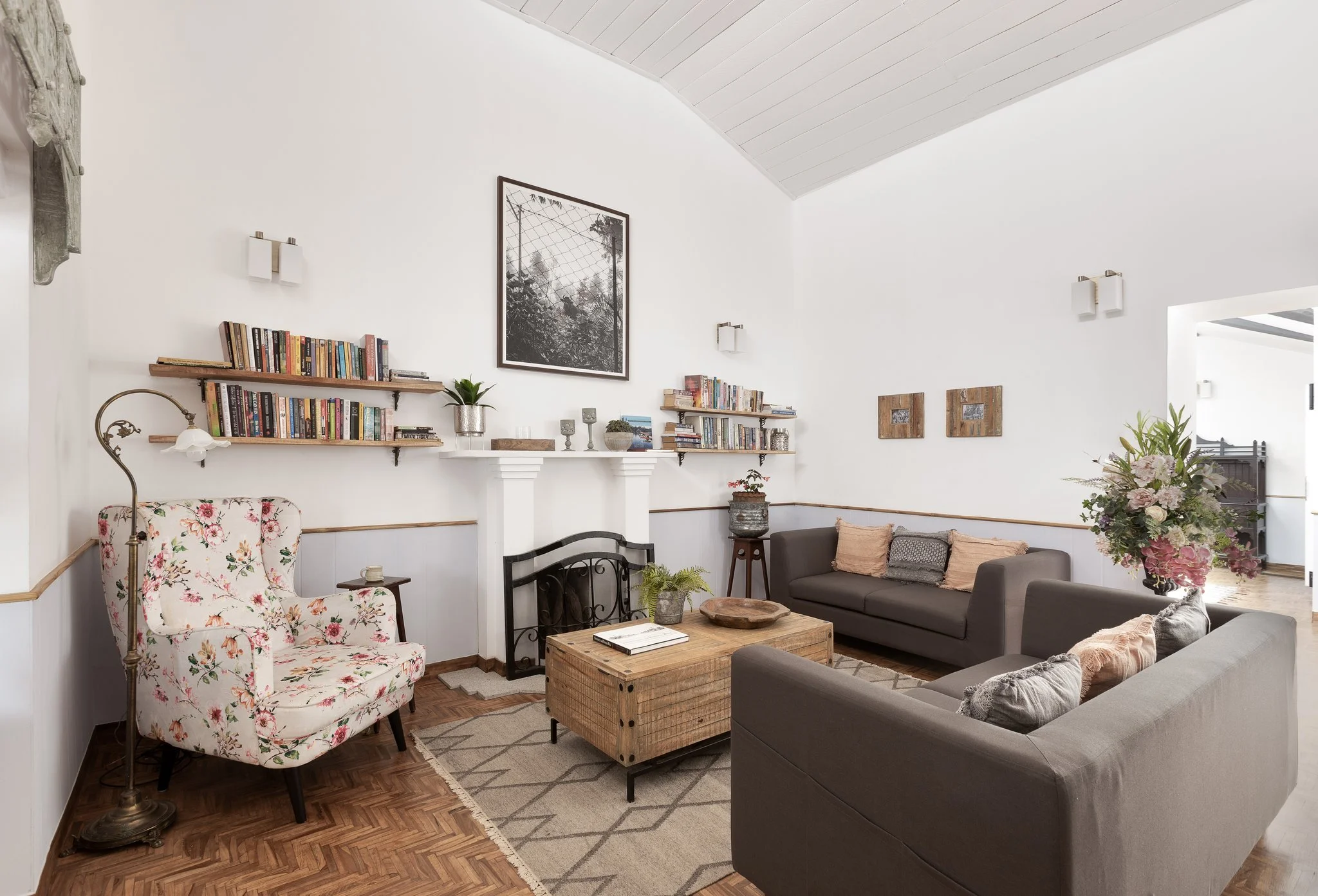 Living room with white walls and wooden flooring, decorated with bookshelves, framed art, sofa, floral armchair, and floral arrangement.