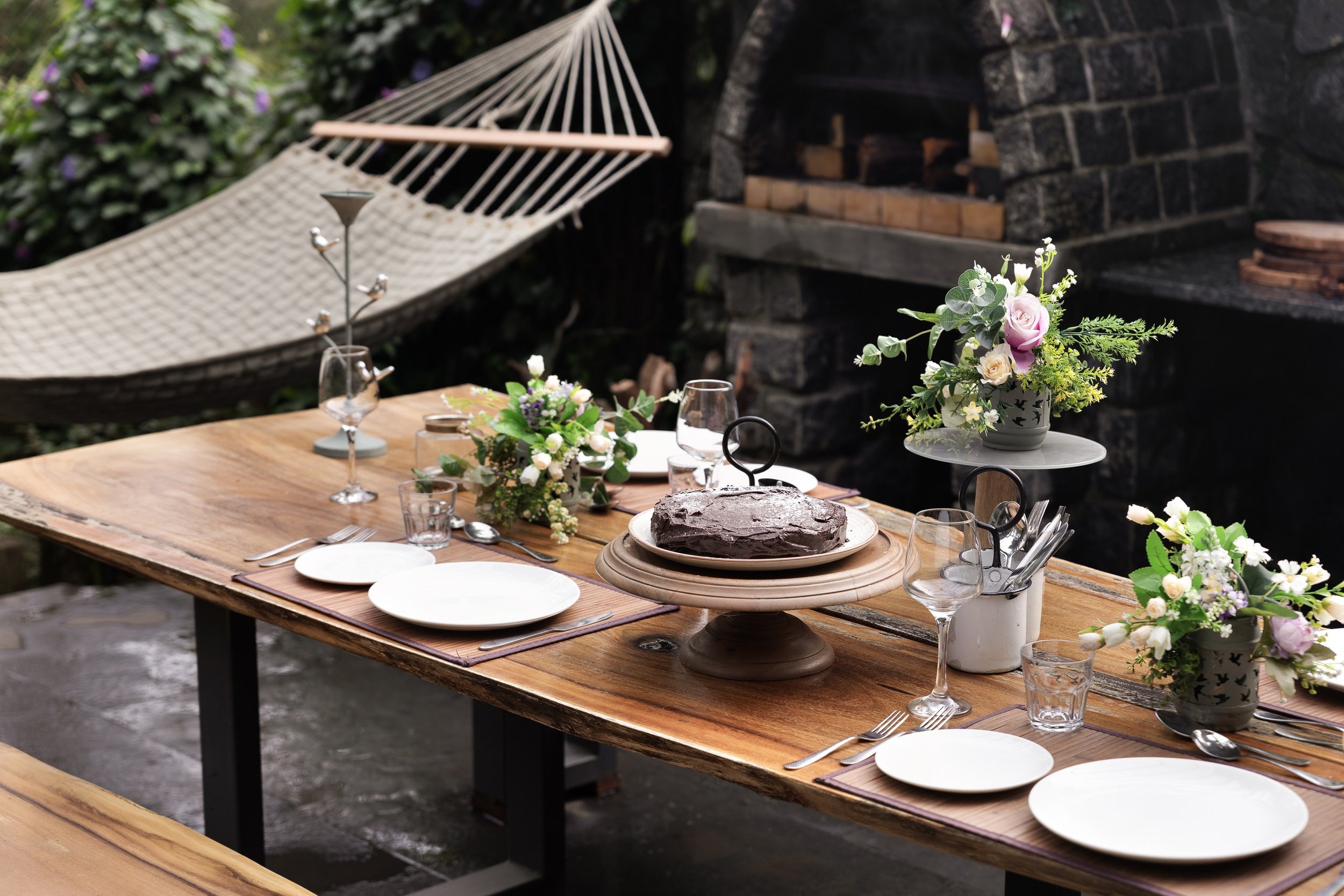 Outdoor dining table set with floral arrangements, glassware, and a chocolate cake, in a garden setting with a hammock and a brick fireplace in the background.