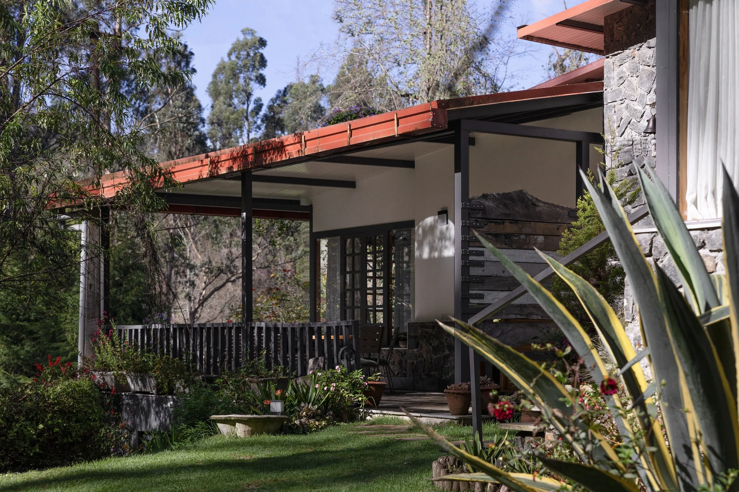 A house with a stone wall exterior, a porch with a railing, and a roof with red tiles, surrounded by various plants and trees.