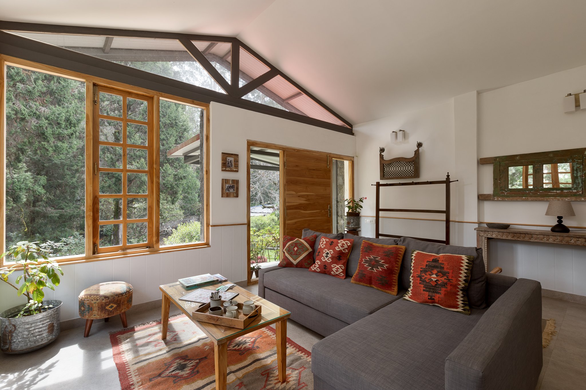 Living room with large windows showcasing forest view, wood accents, and cozy furniture including sofa with patterned pillows, wooden coffee table, and decorative items.