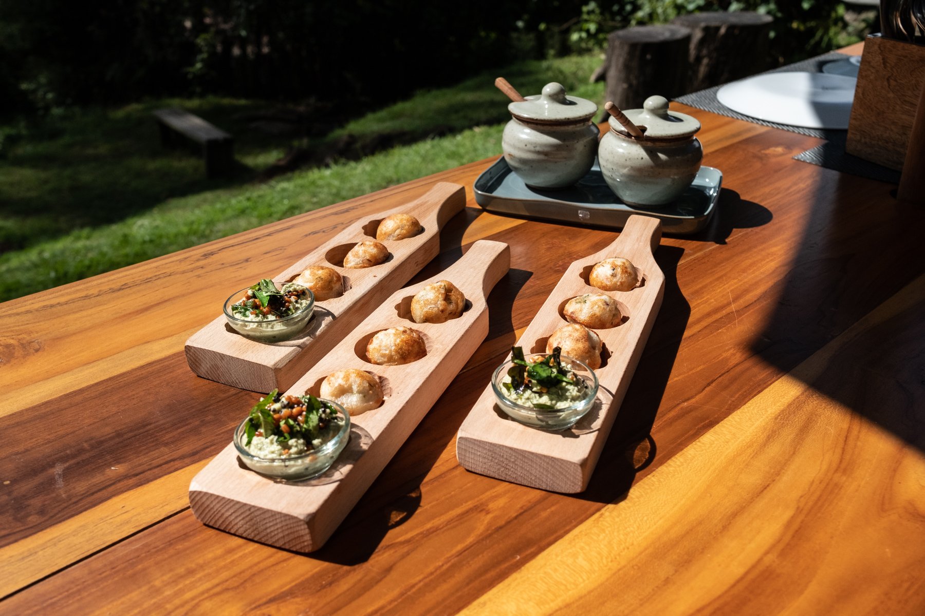 A wooden outdoor table with three wooden boards holding round appetizers, and three small glass bowls with salads, set near garden greenery.