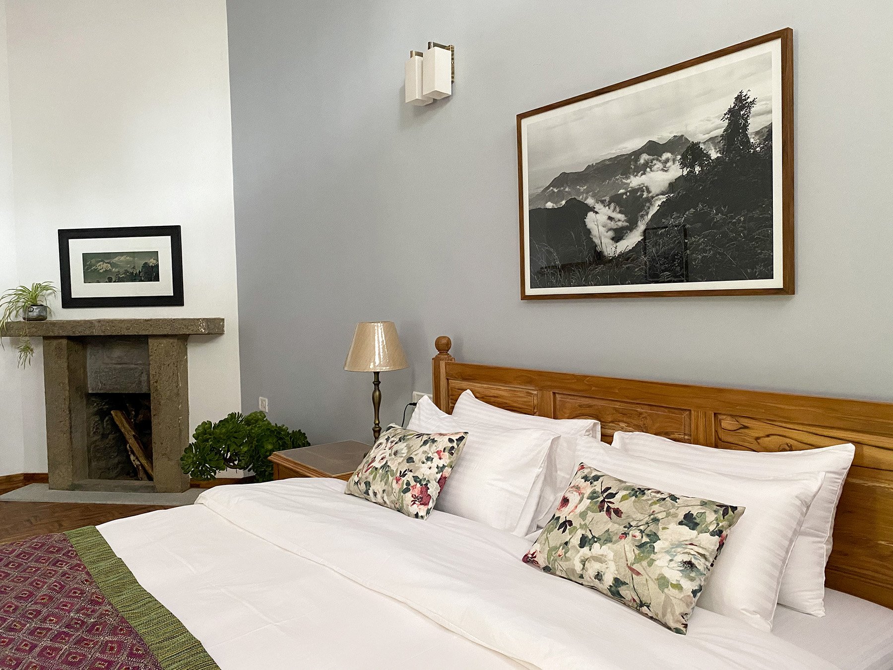 A cozy bedroom with a wooden bed frame, white bedding, and decorative floral pillows. There is a nightstand with a lamp, artwork of mountains and clouds on the wall, and a framed landscape picture above a fireplace with a plant on the mantel.