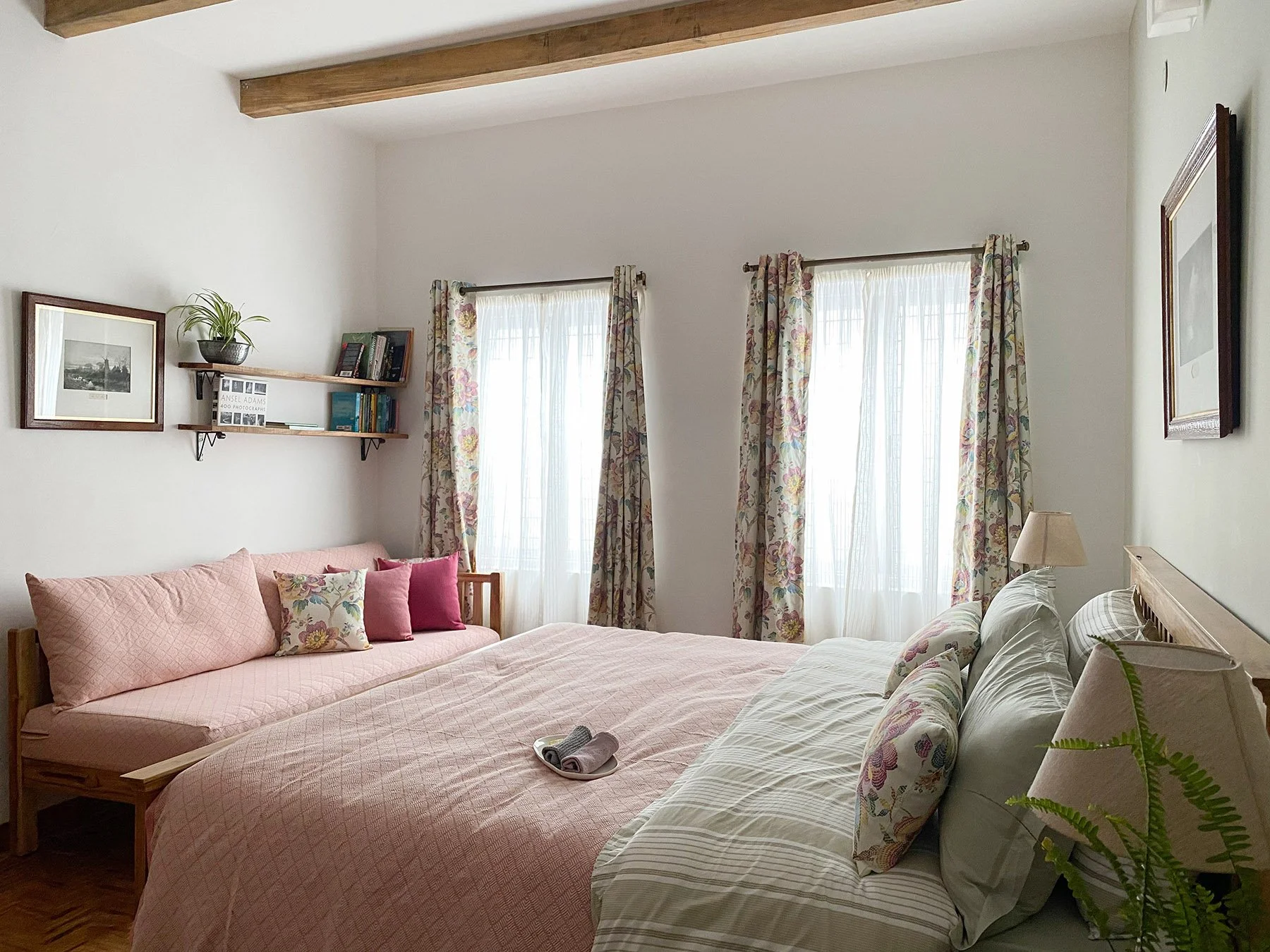 A cozy bedroom with two beds, one with pink bedding and pillows, the other with striped bedding and floral pillows. There are two large windows with floral curtains, a wooden side table with a lamp, framed art on the walls, and a wooden shelf with bo