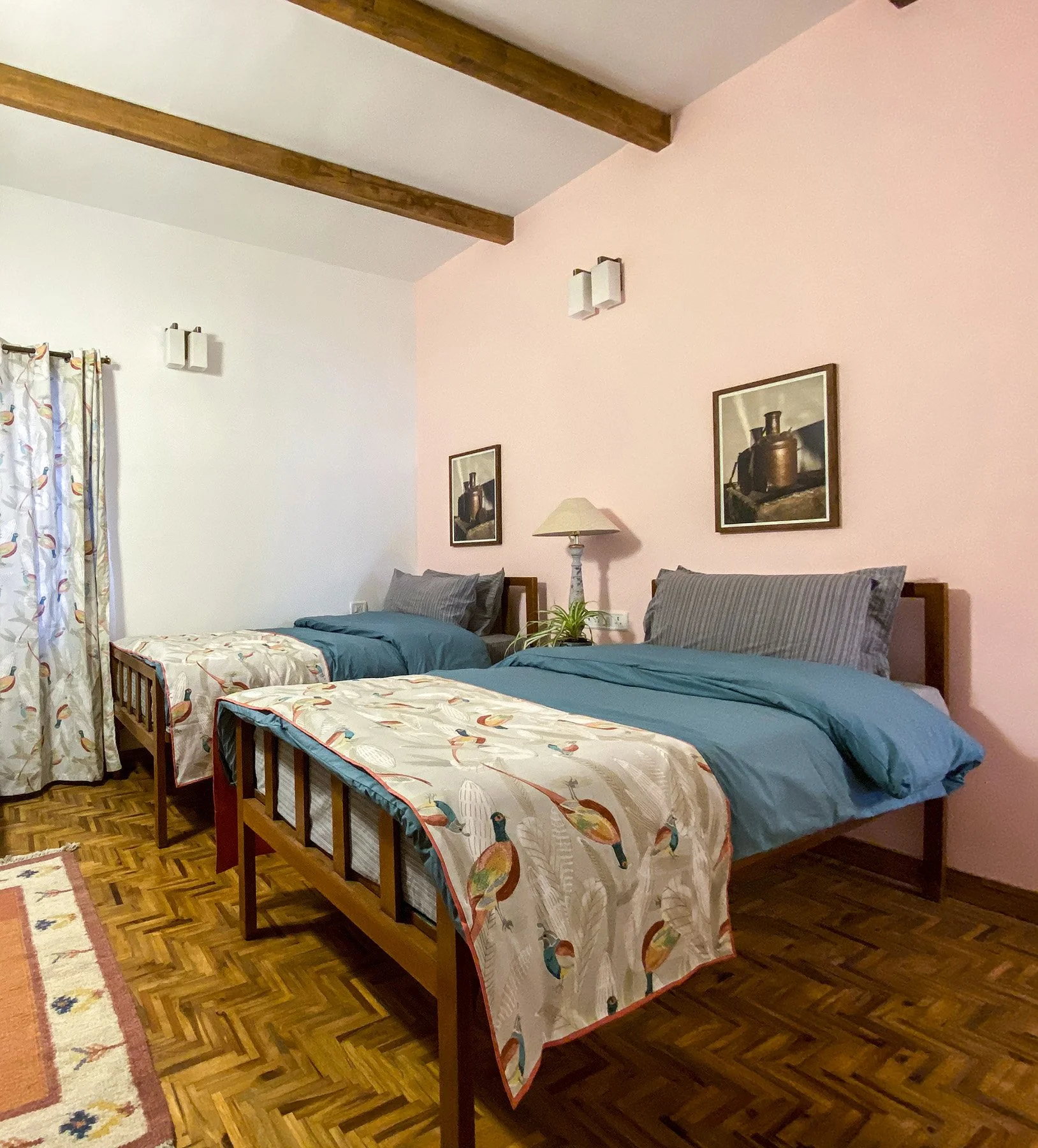 A bedroom with two twin beds with wood frames, decorated with fish-themed bedspreads and striped pillows. The headboard wall has two framed prints of copper kettles, and a pink wall with a lamp and a plant. There are wooden beams on the ceiling, a cu