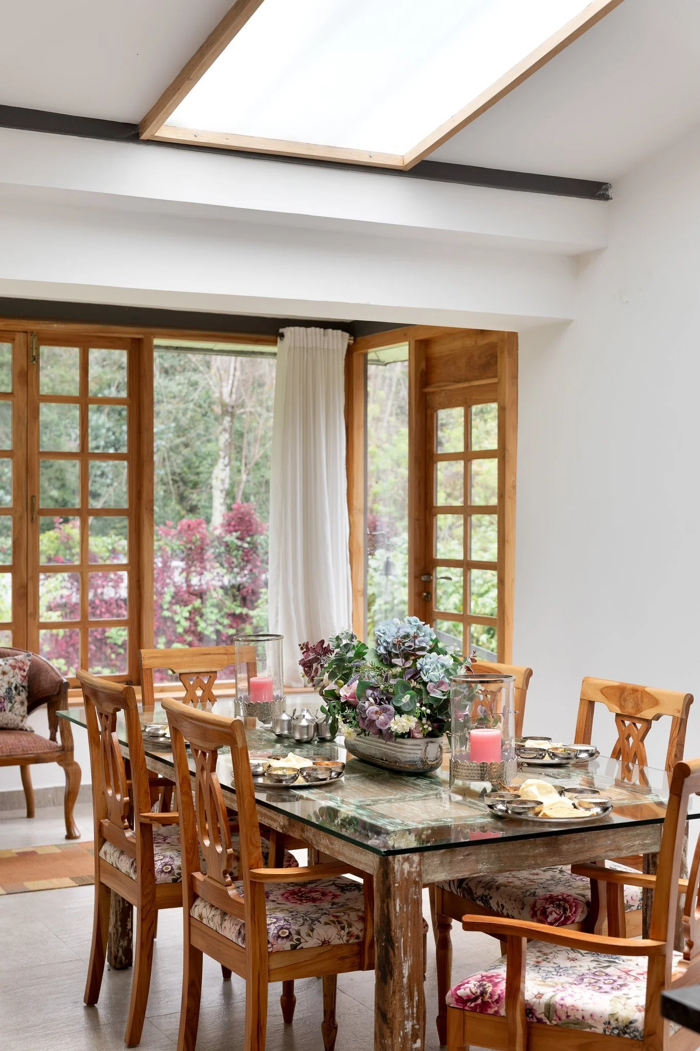 Dining room with a wooden table decorated with a floral centerpiece, surrounded by wooden chairs with floral and patterned cushions. Large windows with wooden frames and white curtains allow natural light to fill the room. The ceiling has a skylight,
