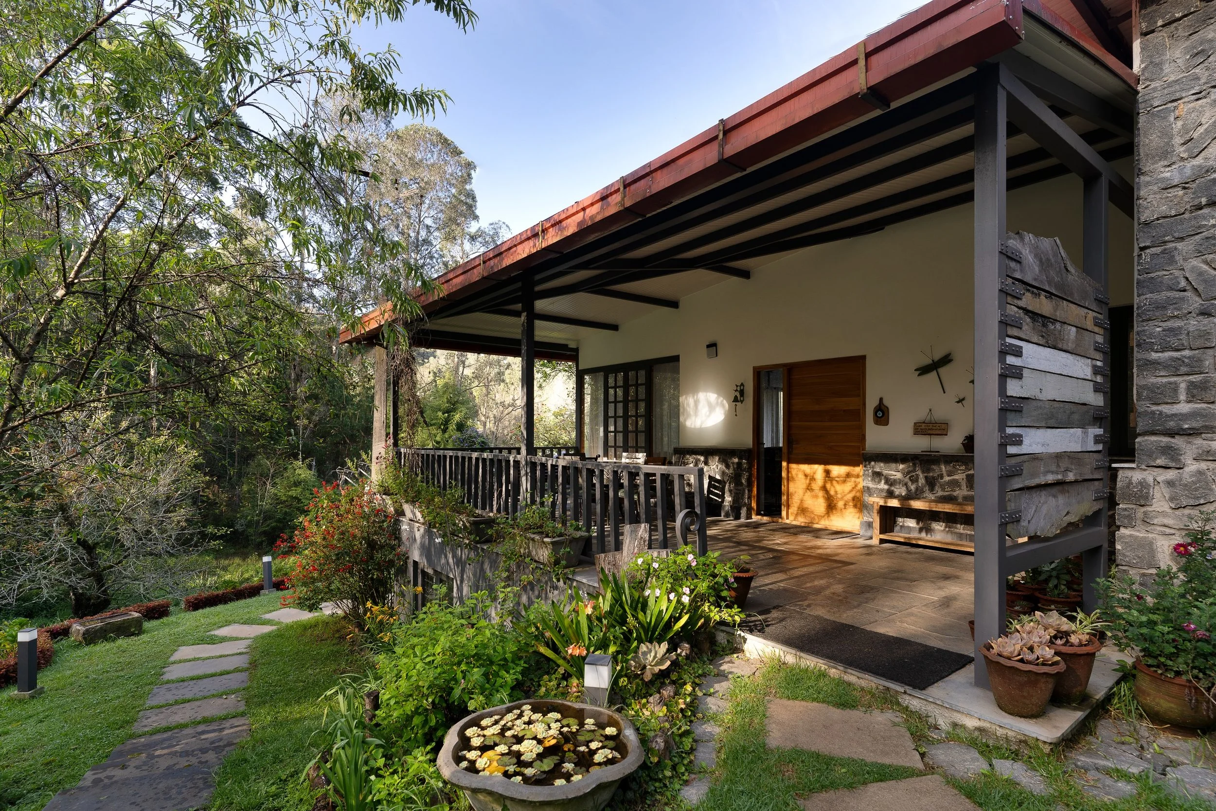 Porch with wooden door and overhanging roof, surrounded by lush garden with potted plants, pathway, and trees.