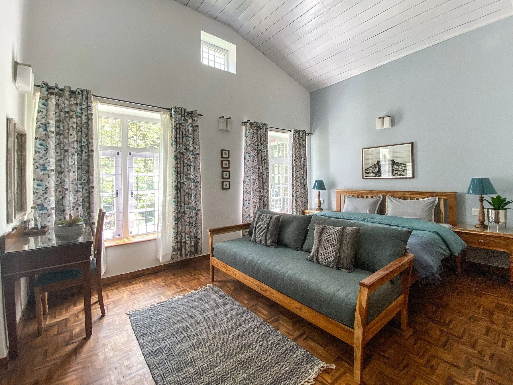 A cozy bedroom with a wooden bed frame and a teal bed spread, decorated with multiple pillows. There are two matching teal lamps on bedside tables. The room has light blue walls, a high vaulted ceiling with white paneling, and windows with floral cur