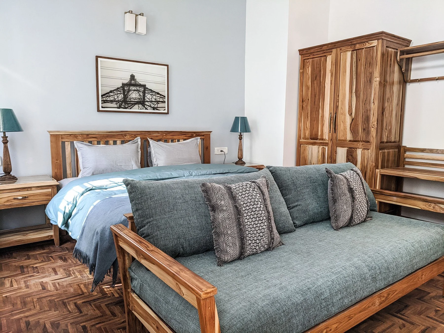 A bedroom with a wooden bed frame, blue and gray bedding, a sofa with patterned cushions, bedside tables with teal lamps, a wooden wardrobe, and a framed photograph of a bridge on the wall.