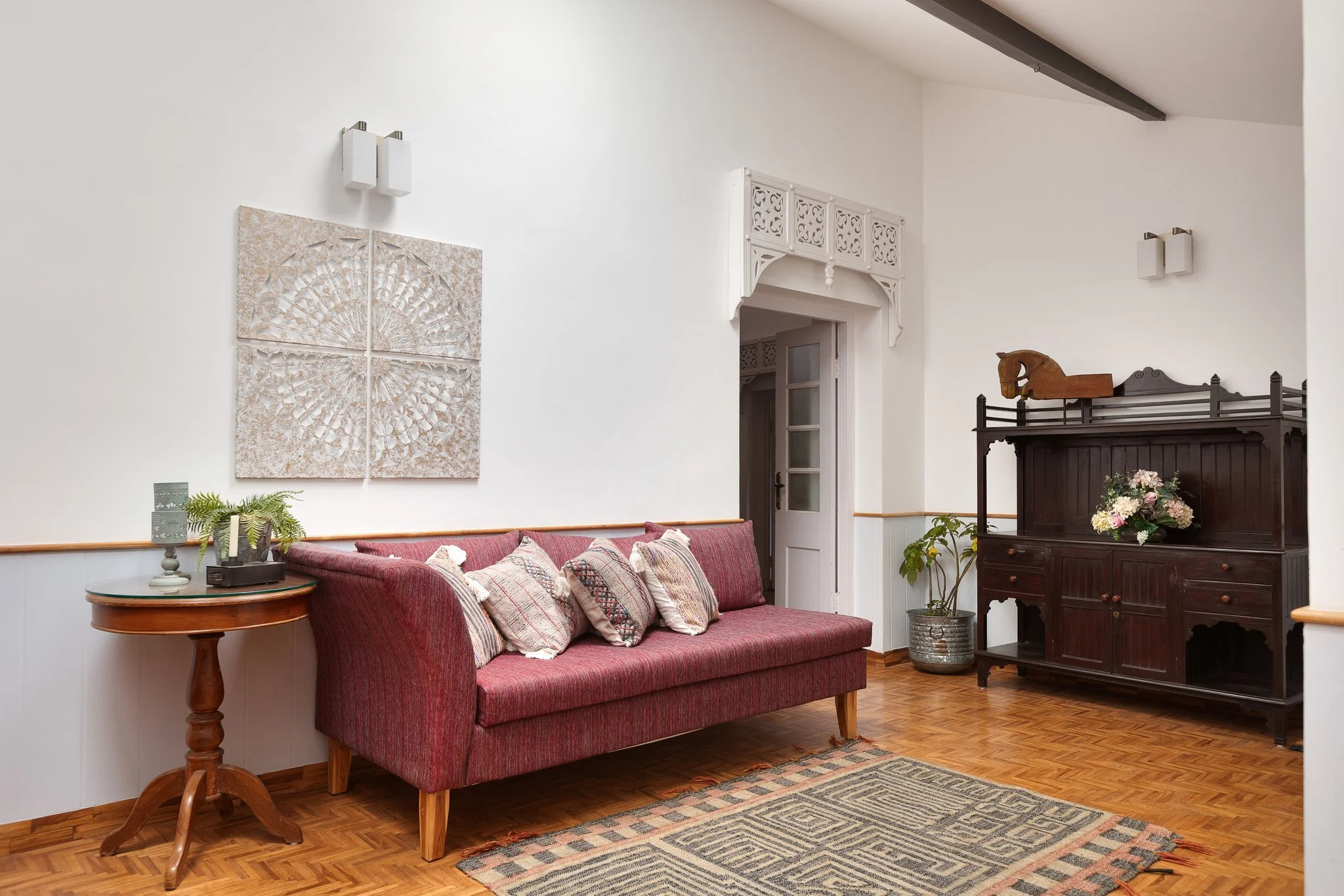 A cozy living room with a red upholstered sofa decorated with patterned throws and pillows, a round wooden side table with a potted plant and decorative items, a dark wooden cabinet with a carved horse head figure and flowers, and wall-mounted lighti