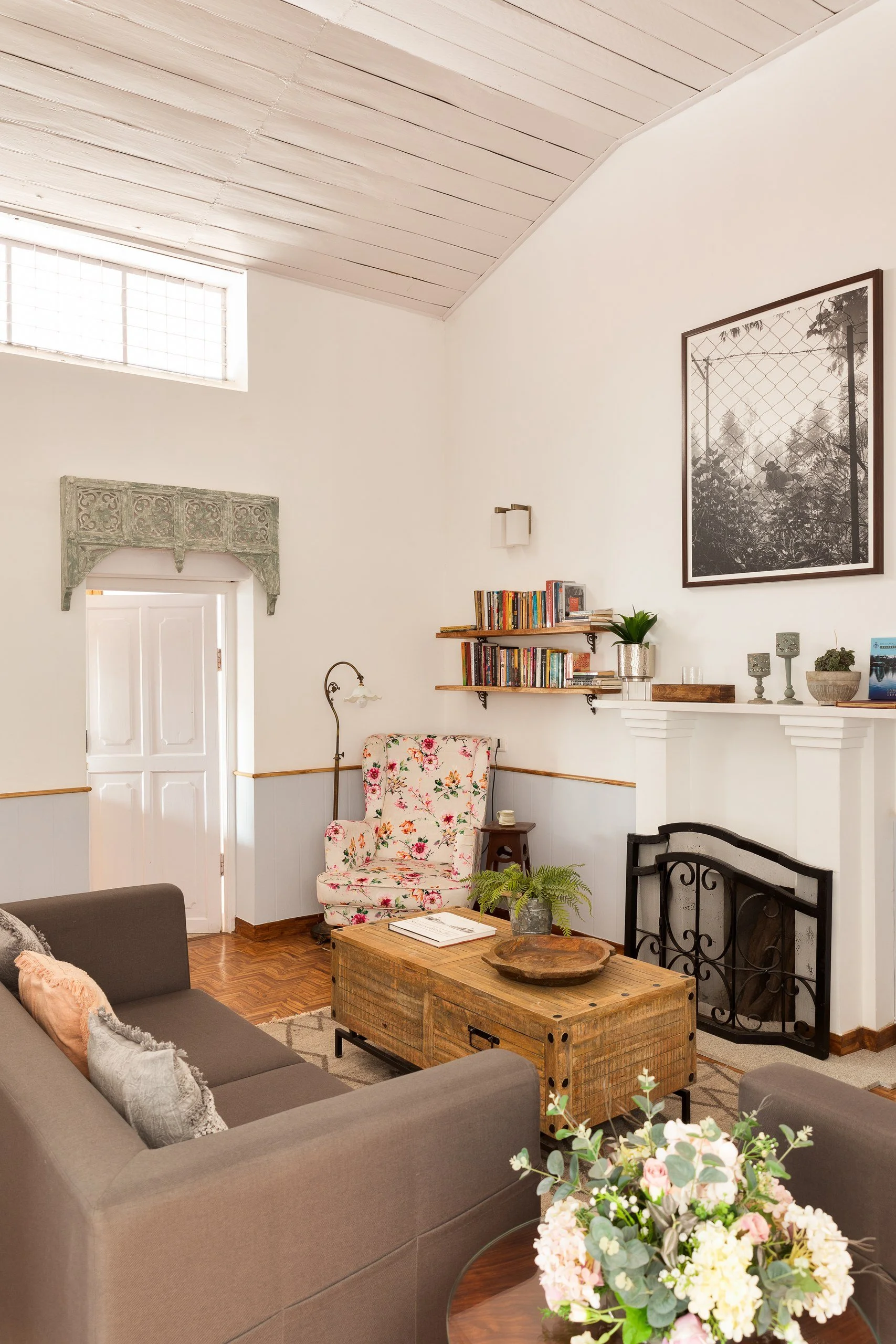 Living room with a gray sofa and floral armchair, wooden coffee table with decorative bowl, bookshelf, fireplace, and artwork on white walls, hardwood floor, and vaulted ceiling.