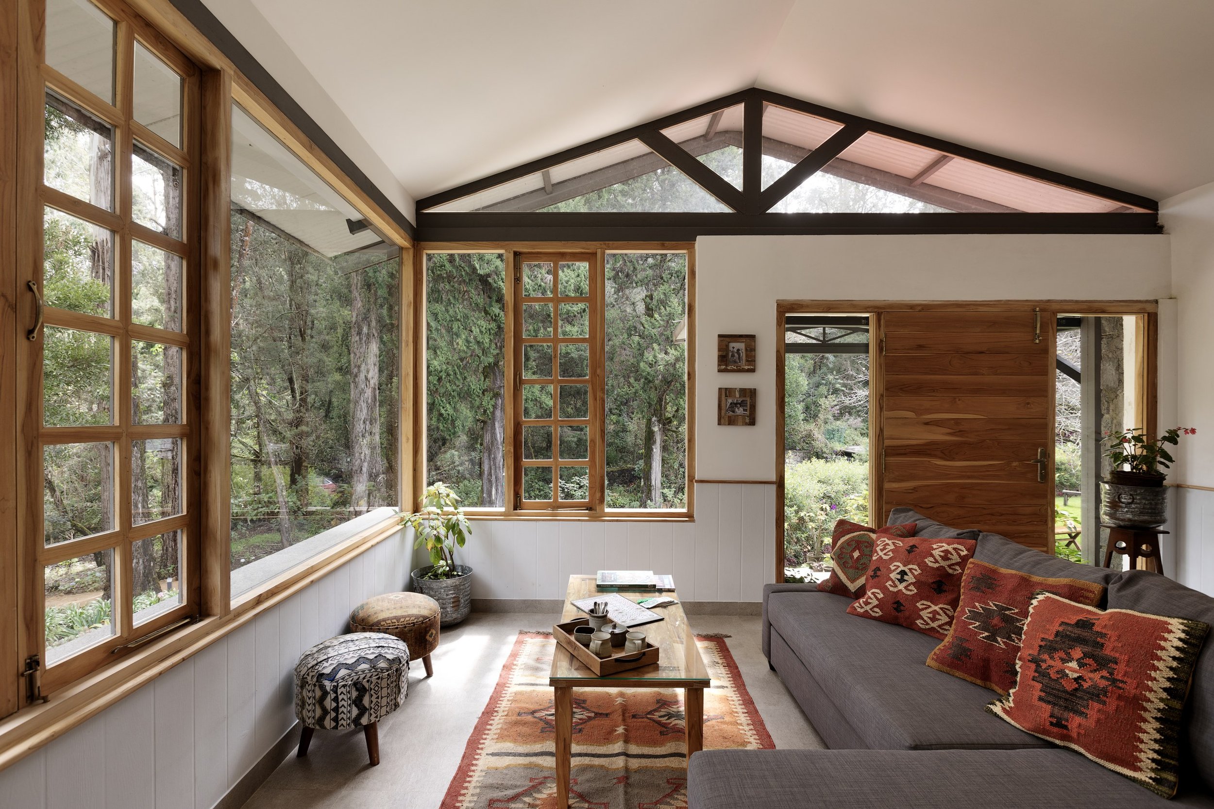 Interior view of a cozy living room with large windows showing a wooded outdoor scene, wooden framing, a gray couch with patterned pillows, a wooden coffee table, two small stools, and plants.