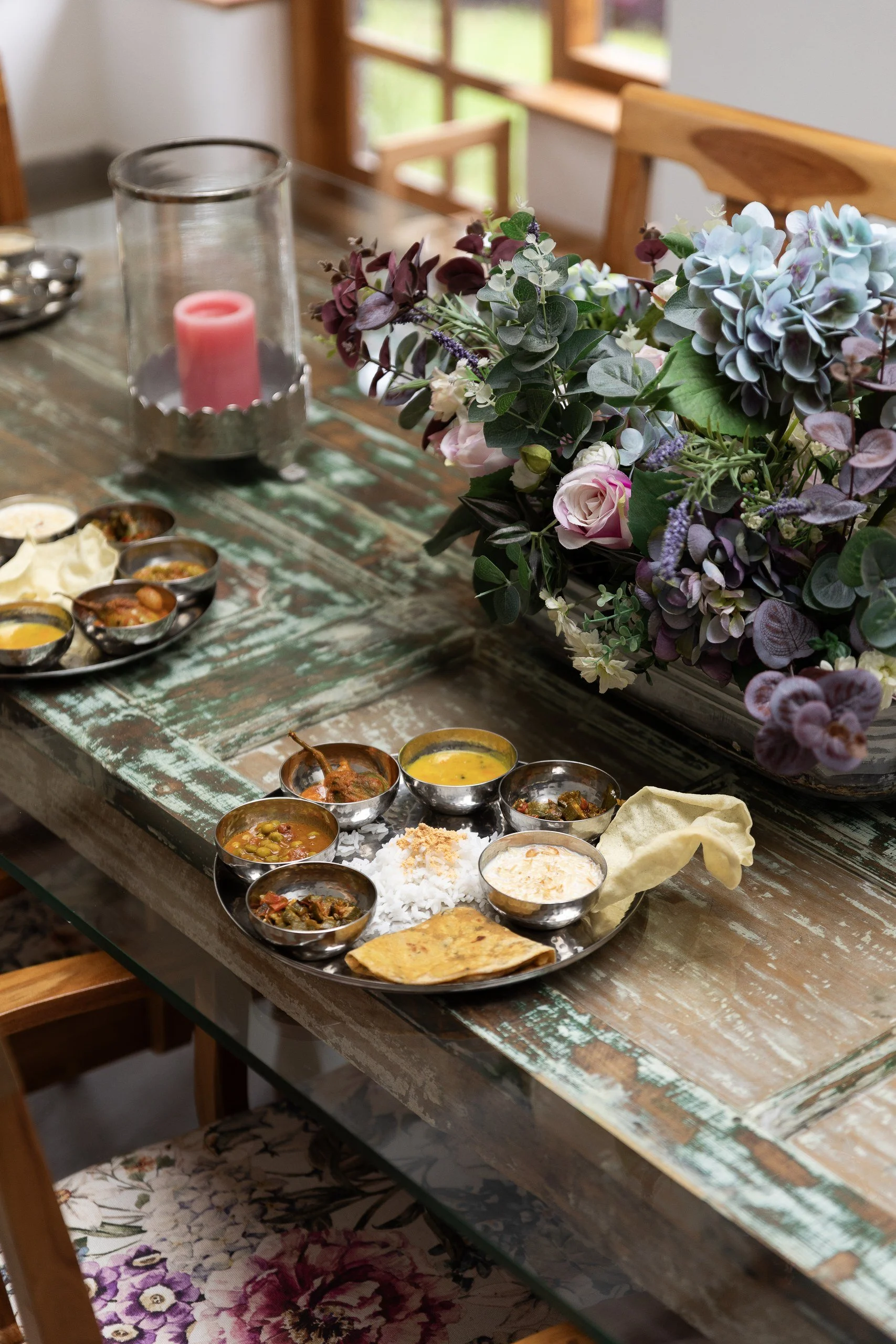 A rustic wooden table with a floral centerpiece, a glass candle holder with a pink candle, and a traditional Indian meal served on a silver tray with small bowls of various curries, rice, bread, and side dishes.