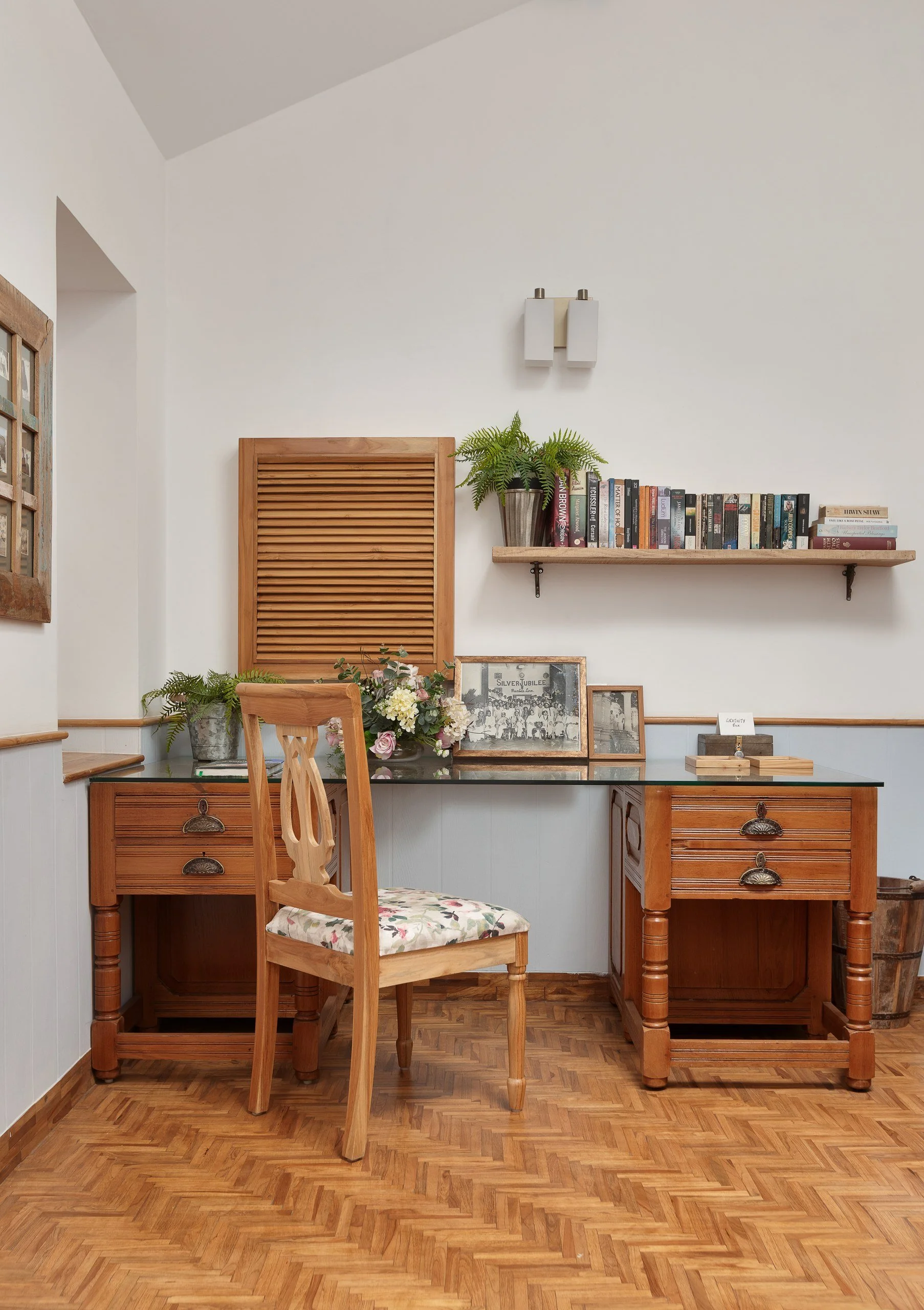 A cozy dining corner with a wooden table and chair, a bed of flowers, a bookshelf, and framed photos on the wall, all with white walls and wooden flooring.