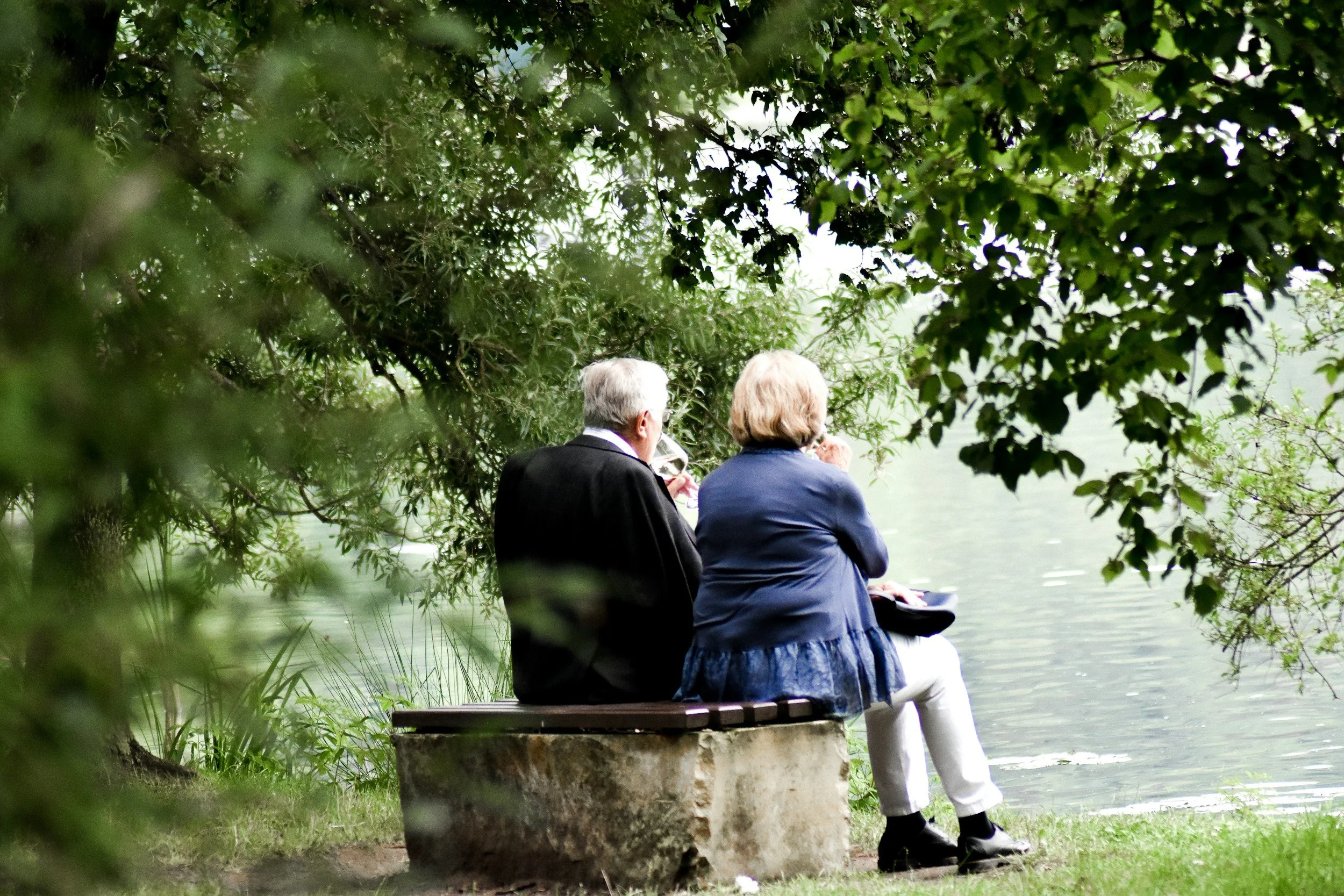 Two elderly people, a man with gray hair and a woman with blonde hair, sit on a bench by a lake, surrounded by green trees and foliage, enjoying drinks.