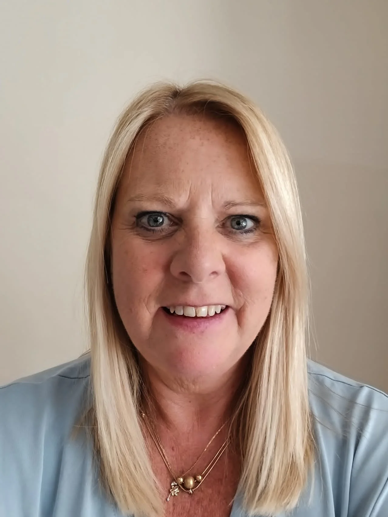 Close-up photo of a middle-aged woman with blonde hair, blue eyes, smiling, wearing jewelry and a blue shirt against a plain background.