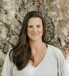 A woman with long brown hair standing in front of a large tree trunk, wearing a white shirt with a gray sweater. She is smiling at the camera.