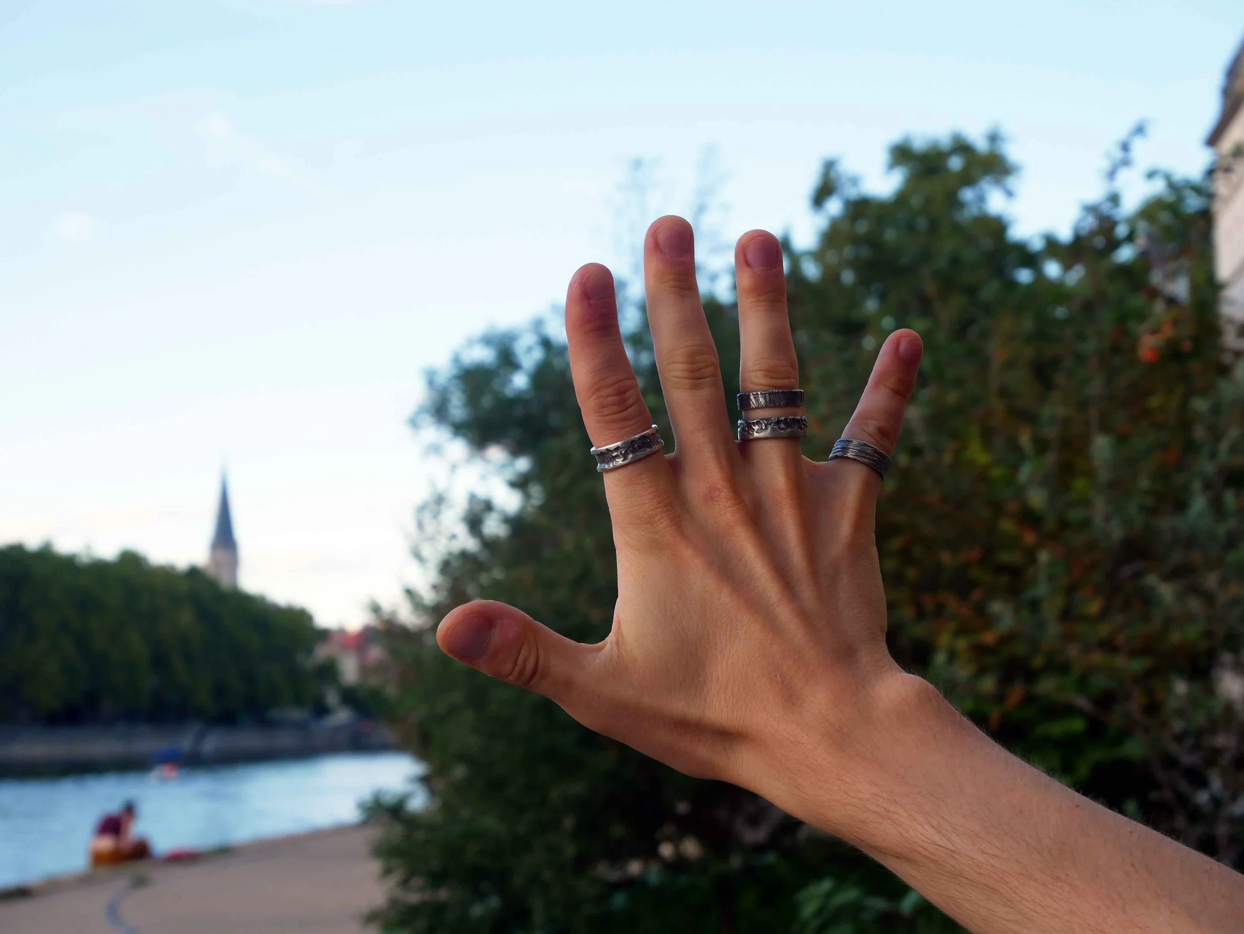 Main d’homme s’étirant face à paysage de Lyon, portant quatre bagues en argent. Bijoux masculins stylés en argent 925 sur fond urbain lyonnais.