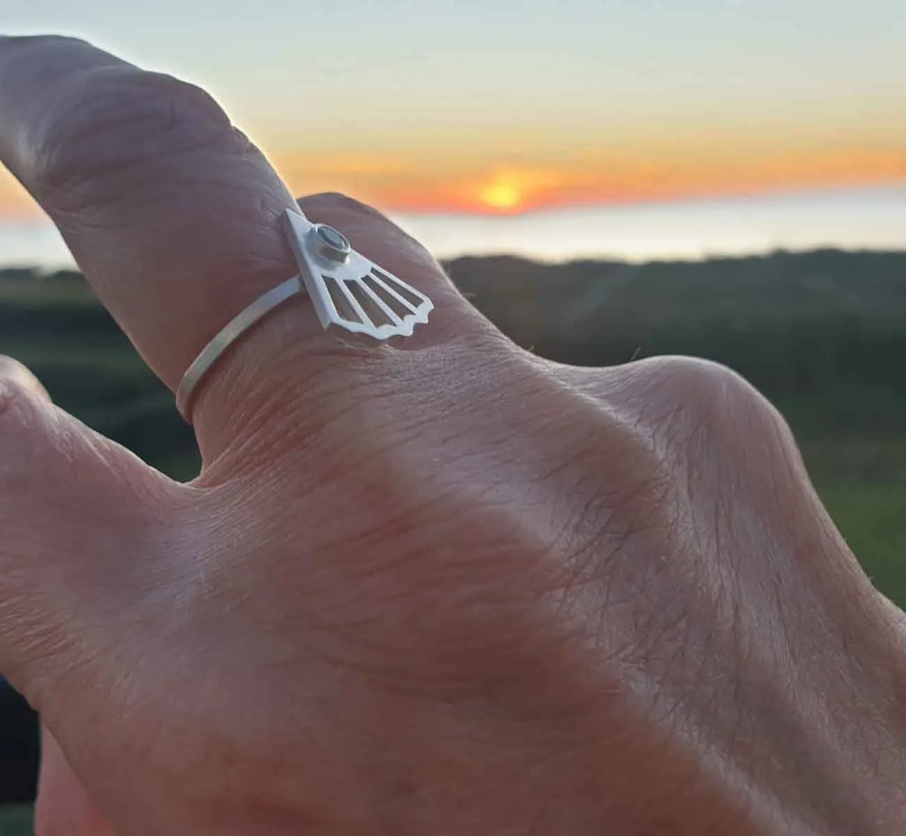Main de femme de 40 ans portant une bague aux formes végétales ajourées sertie d’une pierre verte claire, avec un paysage flou derrière. Bijou élégant texturé réalisé sur-mesure à Lyon.