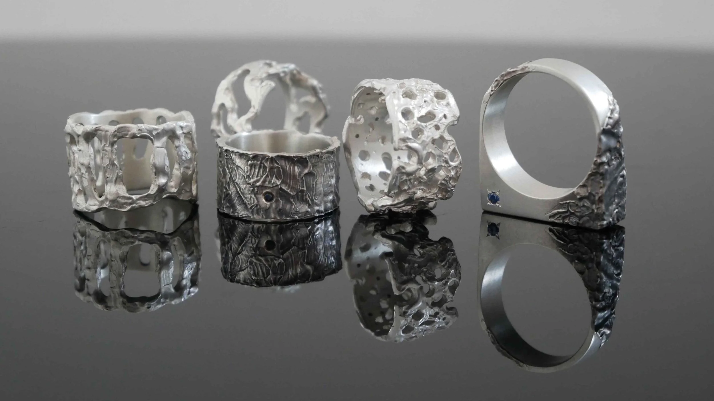 Bagues en argent avec pierres précieuses sur surface noire réfléchissante : larges ajourées, avec gemmes, saphir. Reflets captivants mettent en valeur ces bagues en argent avec des pierres précieuses.