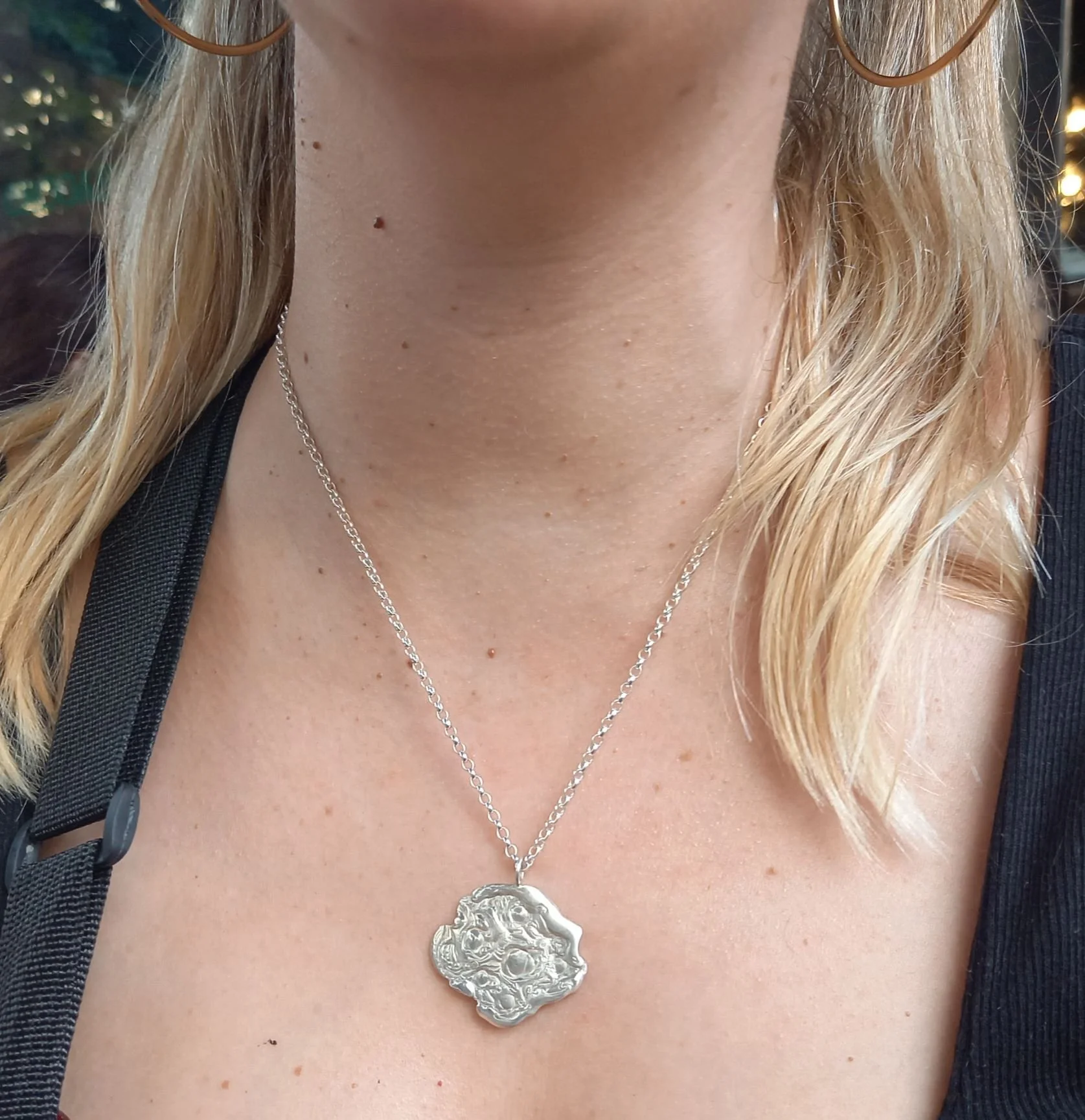 Plan serré sur le cou d’une femme où pend un collier en argent aux reflets subtils et texture travaillée. Bijou contemporain d’une créatrice à Lyon, il joue sur finition mate pour un effet poétique au quotidien.