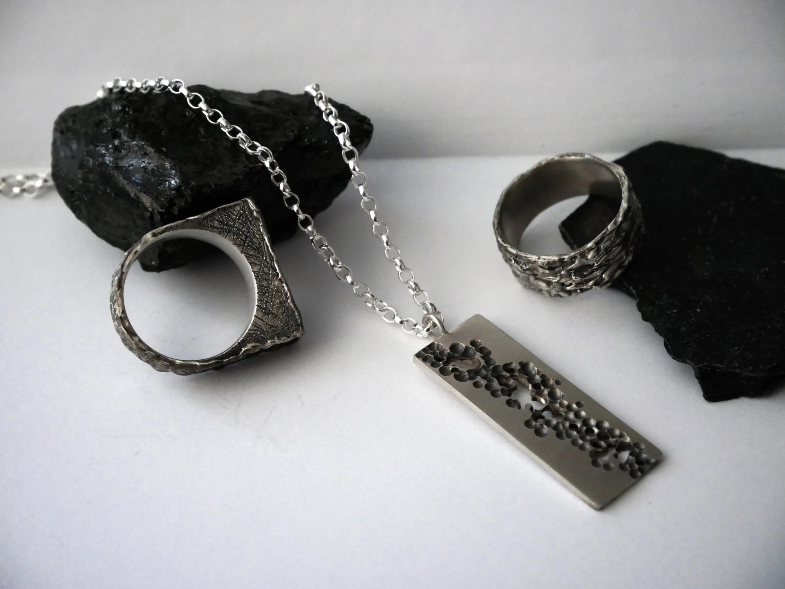 Deux bagues en argent massif et un collier ajouré en argent posés sur fond blanc avec deux cailloux noirs. Bijoux artisanaux aux textures raffinées, idéaux pour femme ou homme amateurs de bague en argent originale et bague argent 925.