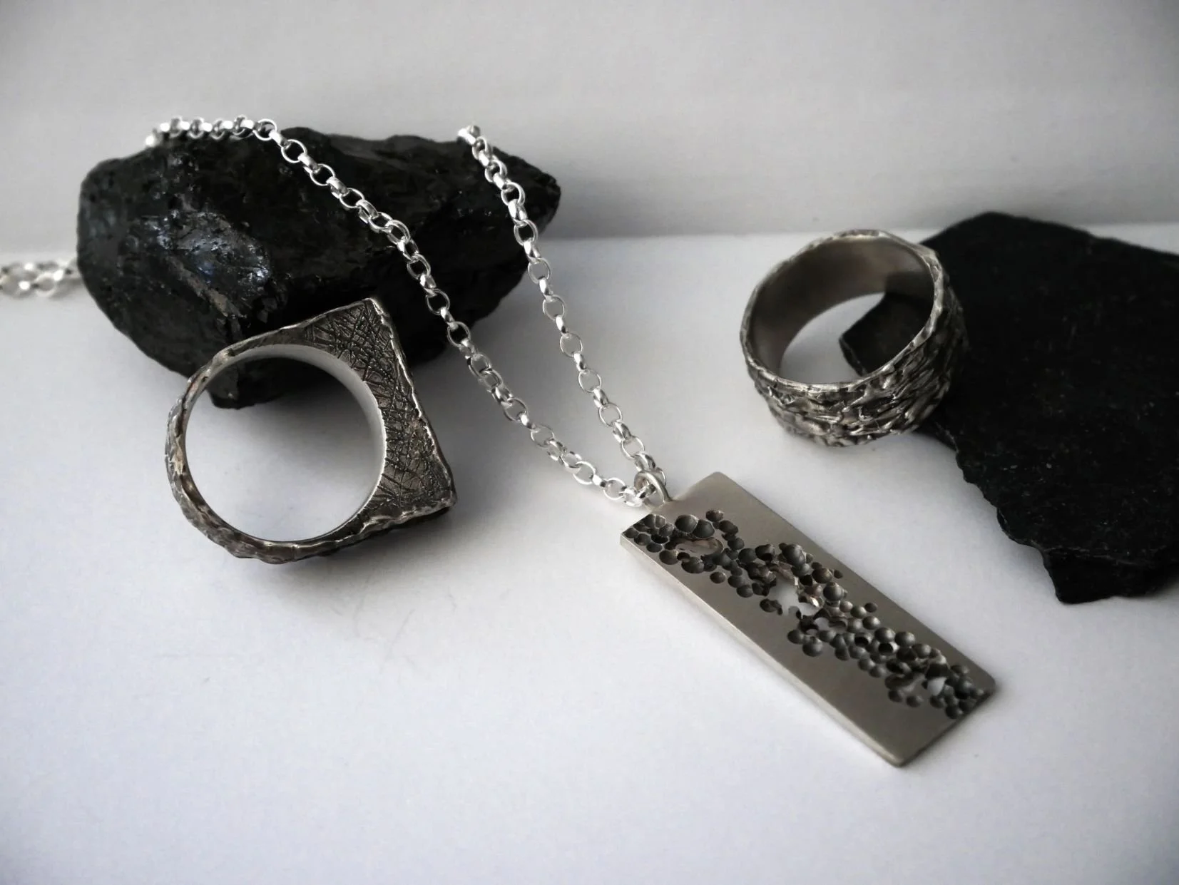 Trois bijoux modernes posés sur des pierres noires et sur un fond blanc. Il y a un collier ajouré et deux bagues : une carrée et un anneau texturé.