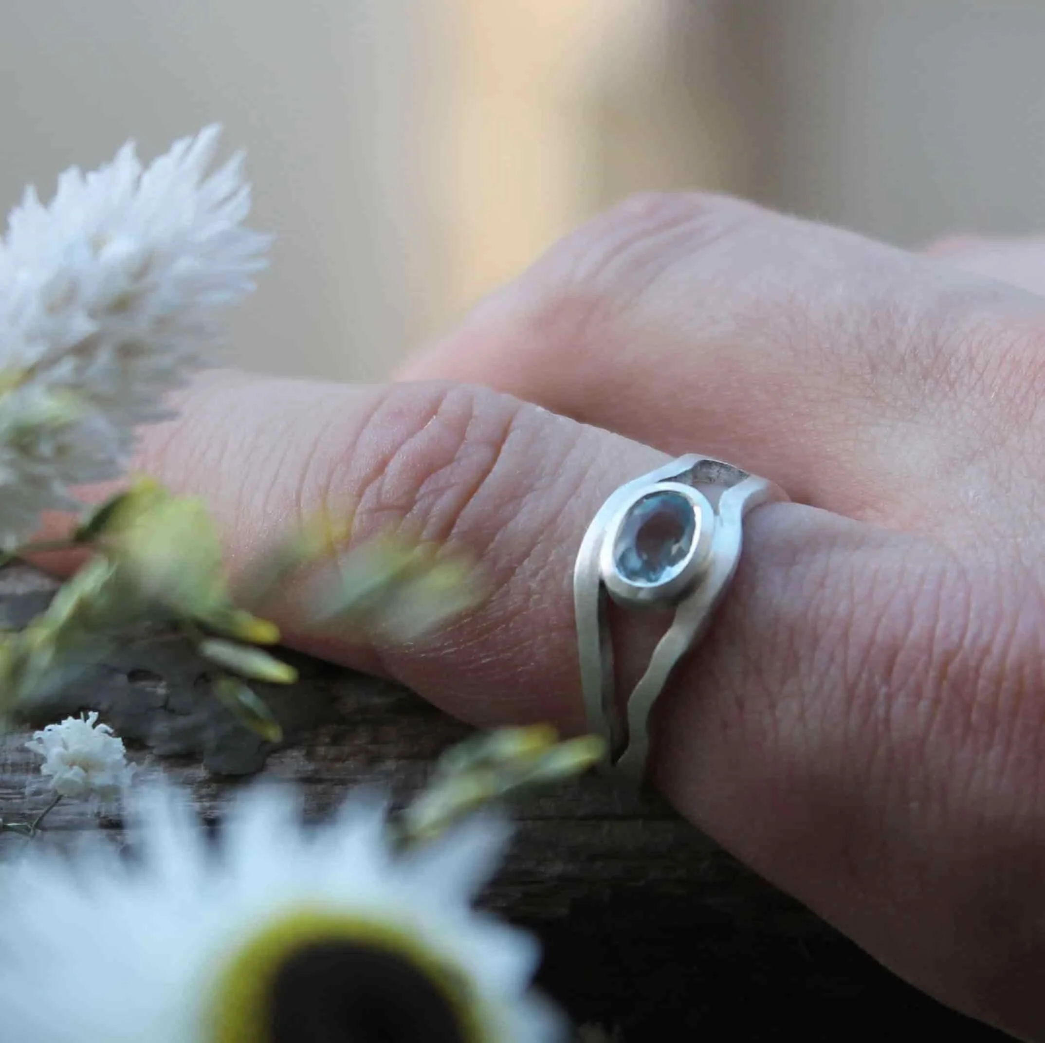 Bague aigue marine moderne sur-mesure, en argent ajouré, au design original. Sertie d’une pierre ovale bleu clair translucide, elle se dévoile portée au doigt parmi des fleurs, symbole de finesse et d’élégance artisanale.