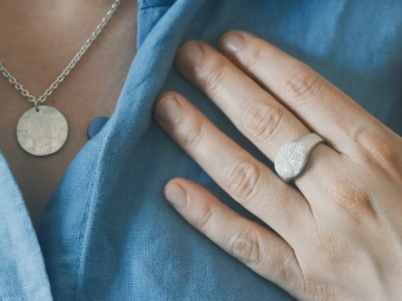 Plusieurs bijoux empreinte de chien sont portés par une femme : une chevalière truffe portée à la main et un collier truffe porté au cou. La femme porte une chemise bleue.