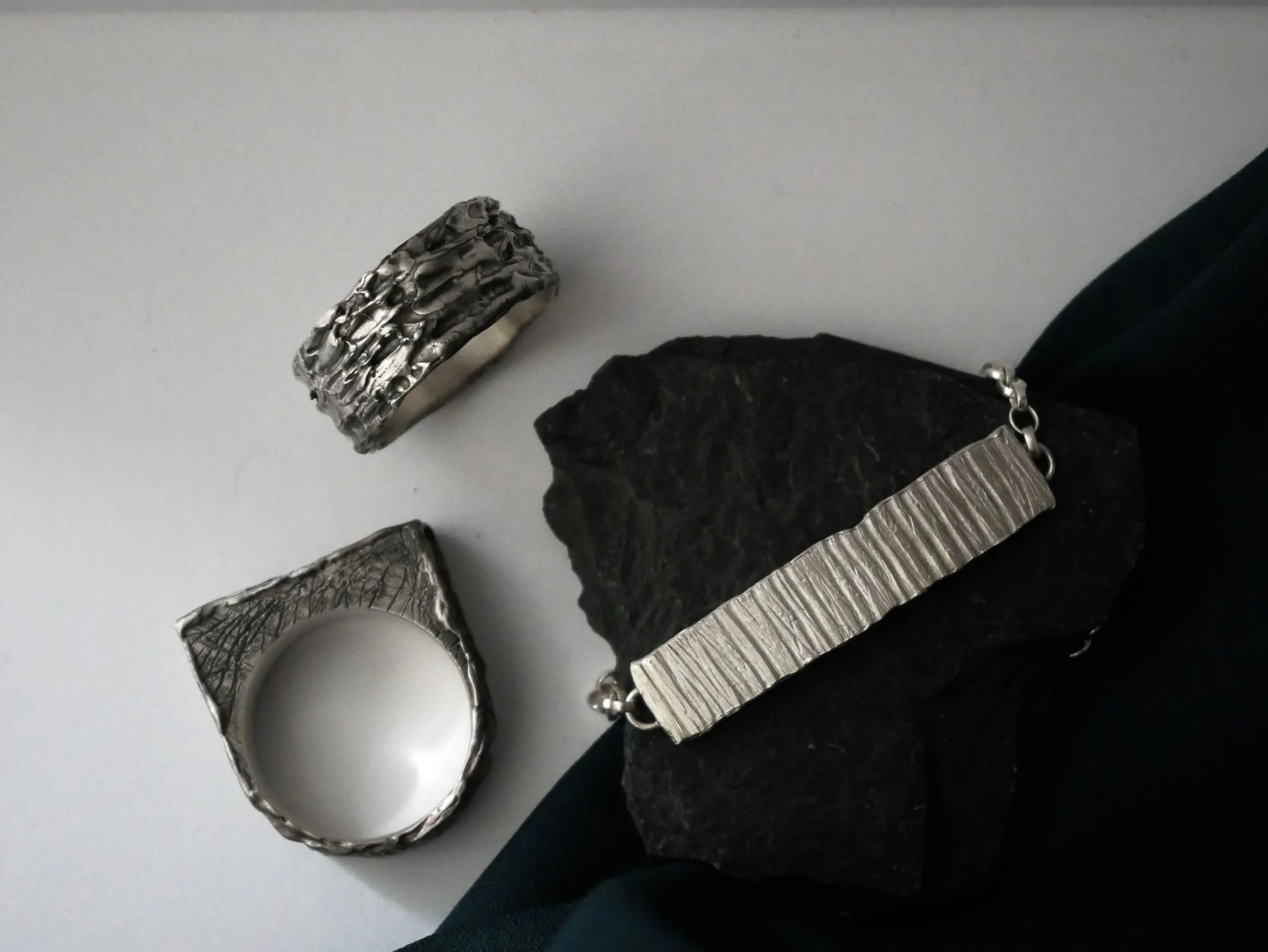 Bijoux en argent : bracelet gourmette texturé, bague carrée et anneau, bracelet sur caillou noir fond blanc. Ensemble mixte en argent massif.