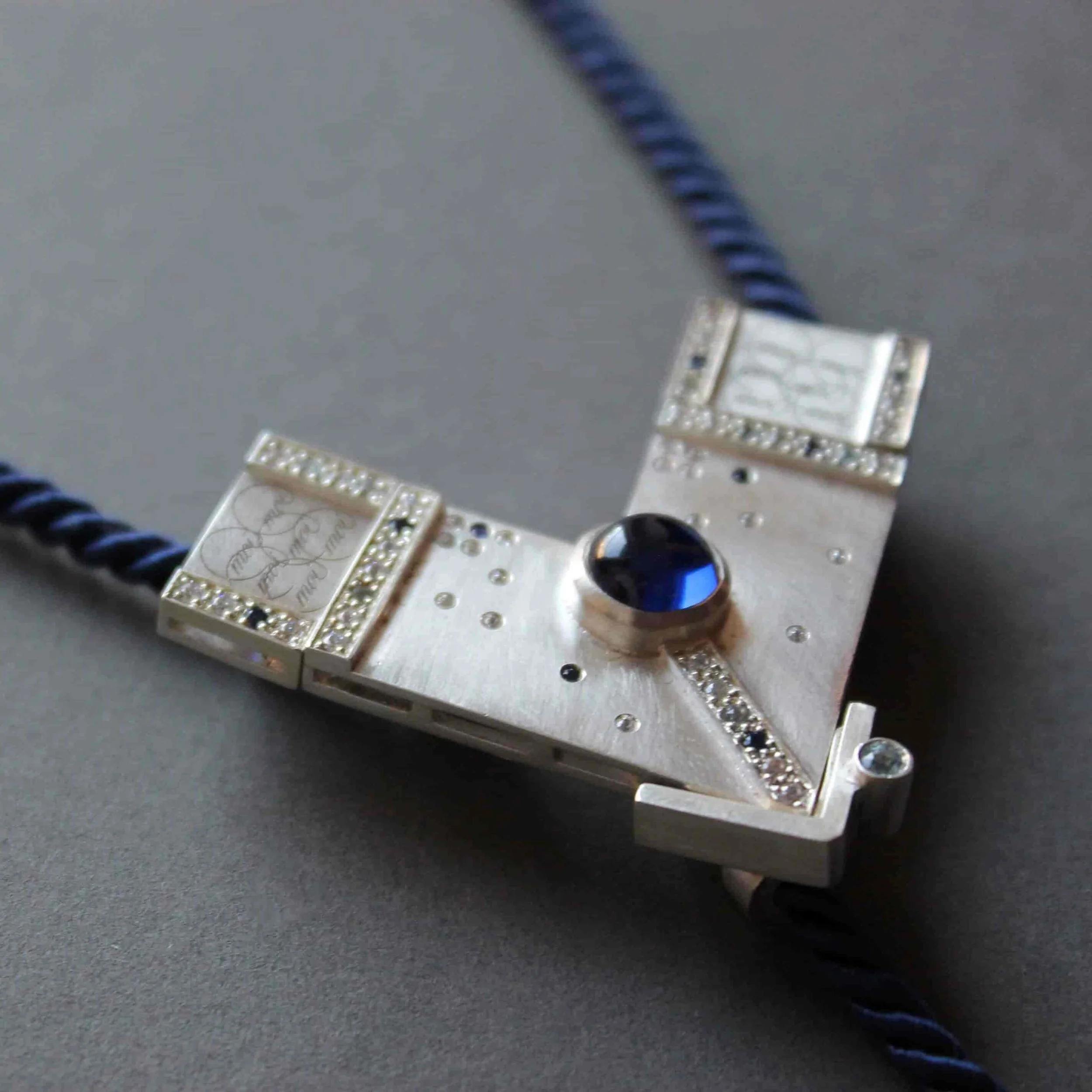 Sautoir femme en argent sur-mesure, modulable et serti d’un saphir. Son pendentif triangulaire gravé d’inscriptions énigmatiques révèle un bijou unique, à l’esthétique contemporaine et au symbolisme mystérieux.