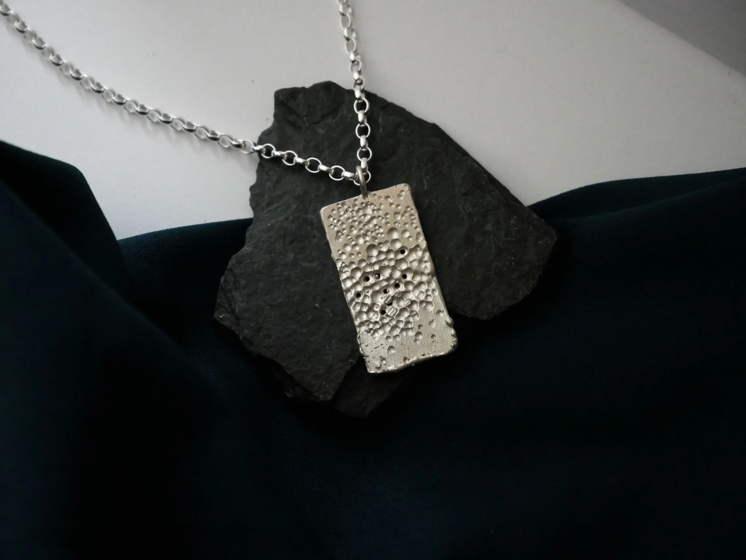 Collier rectangulaire en argent posé sur un caillou noir, fond blanc épuré. Pièce ajourée et contemporaine, issue d’une création artisanale en argent 925.